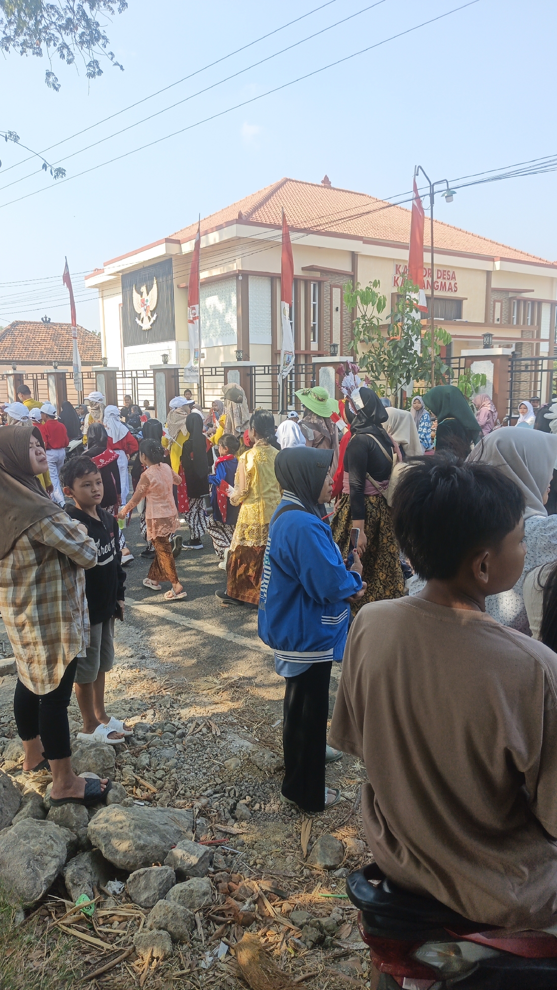The image captures an outdoor gathering of people, likely a local event or celebration, in front of a building that appears to be a village hall or administrative office. The building has a tan facade, a red tiled roof, and is decorated with Indonesian flags. A prominent emblem resembling a Garuda bird is displayed on the building.
A diverse group of individuals, including adults and children, are present. Many are wearing traditional Indonesian attire, such as sarongs and patterned tops. Some children are in brightly colored costumes, suggesting a parade or performance. The ground is a mixture of asphalt and dirt, with rocks and dry leaves scattered around. In the foreground, several people are standing and looking towards the front, while others are in the background, interacting with each other. A motorcycle is visible in the lower right corner. The overall atmosphere appears lively and community-oriented, under a clear blue sky.