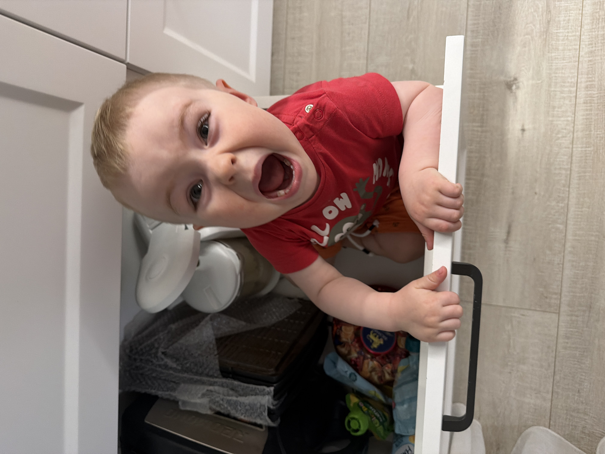 A vibrant young child with light hair and blue-gray eyes is playfully peeking out from an open lower cabinet drawer in a home, likely in Votkinsk, Russia. The child, wearing a red short-sleeved t-shirt with the visible partial text "LLOW" and some indistinct cartoon-like patterns, has a wide-open mouth revealing small teeth, and a surprised or excited expression.
The child's hands are gripping the top edge of the white drawer, which features a black, horizontal handle. Inside the drawer, various items are stored, including white plastic objects (possibly baby shoes or parts of a potty training seat), a grey mesh material, a dark rectangular object, a colorful patterned fabric item, and a small green toy car.
The background shows modern, light-colored flat-panel cabinets to the left and above, suggesting a kitchen or bathroom setting. The flooring is a light wood-look laminate or tile. The scene is brightly lit, indicating daytime, capturing a candid moment of childhood curiosity and exploration within a clean, contemporary home interior.