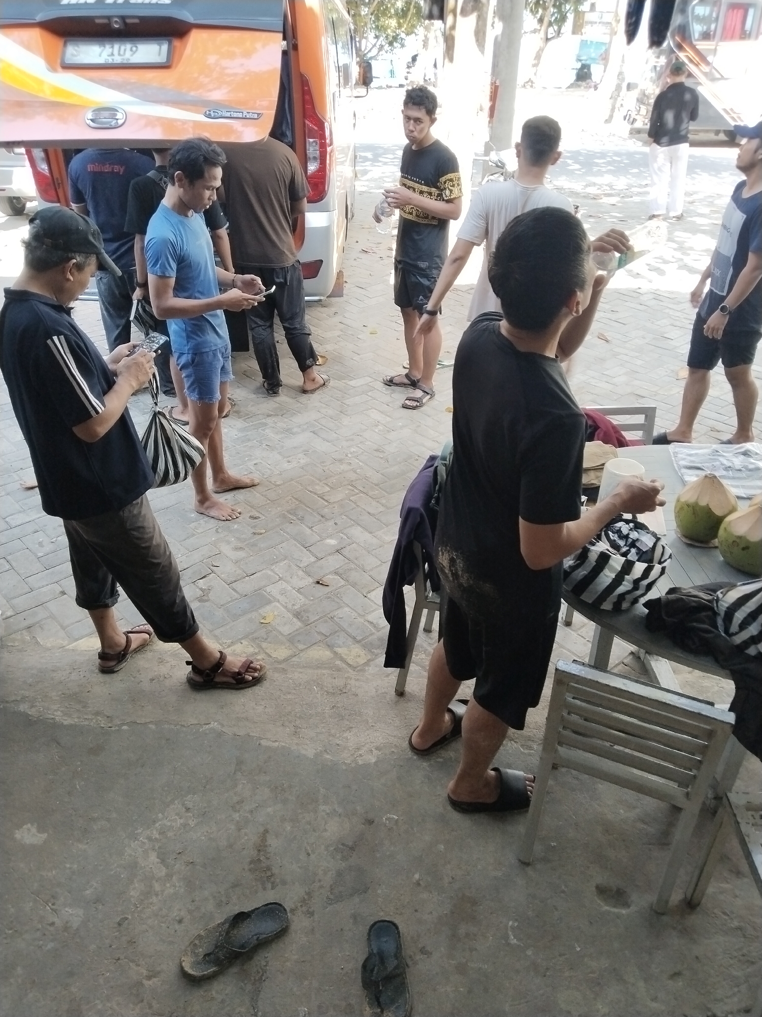 In a street scene in Prigi, Indonesia, a group of individuals are gathered near a transport vehicle, likely a bus, based on the presence of a rear tailgate and the vehicle's overall shape. The ground is paved with interlocking bricks and concrete.
On the left side of the image, a person wearing a black t-shirt, dark pants, and brown sandals is holding a smartphone. Next to them, another individual in a blue t-shirt and denim shorts stands barefoot, also holding a smartphone. Further back, near the vehicle, a person in a brown t-shirt and dark pants is partially visible.
In the center and right of the image, a person wearing a black t-shirt and black shorts is standing at a table. They are holding a white cup and appear to be interacting with items on the table, which include coconuts and a striped bag. Behind them, a person in a white t-shirt and dark shorts is visible. To the far right, a person in a blue t-shirt and dark shorts stands with a water bottle.
Scattered on the ground are several pairs of footwear, including sandals and flip-flops, suggesting a casual environment where people may have removed their shoes. The background shows trees and hints of other structures, characteristic of an outdoor setting in a town or city. The overall atmosphere suggests a pause or gathering during travel or an outing.