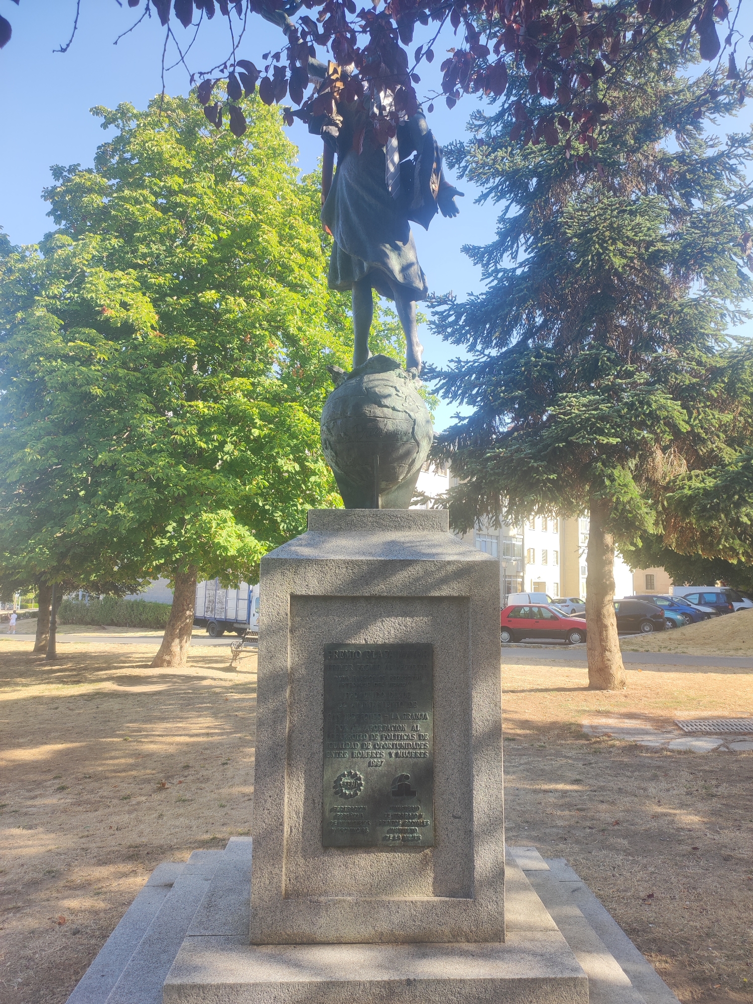 A statue of a person standing on a globe is displayed in a park in Real Sitio de San Ildefonso, Spain. The statue, made of dark metal, depicts the figure with one arm extended forward. The base of the statue is made of stone and features a plaque with text and a small embossed image of a woman. The park setting is characterized by green trees, dry grass, and a paved pathway. In the background, several cars are parked, and a building with windows is visible. The sky is clear and blue.