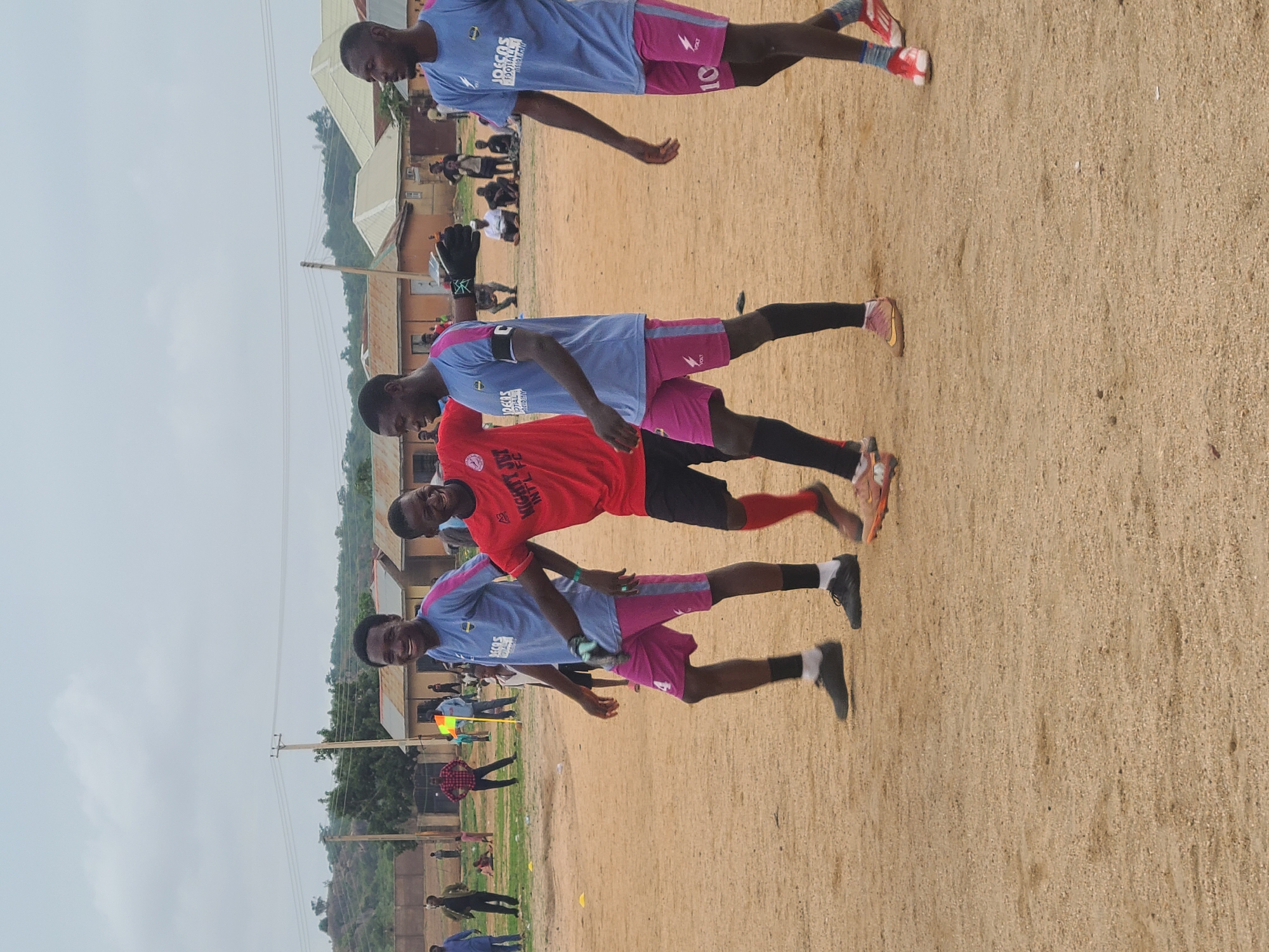The image depicts a group of four men standing on a dirt field in Unknown, Unknown. They are wearing soccer uniforms, with three in light blue and pink, and one in red. The men appear to be posing for the photo, as they are smiling and looking at the camera. One of the men in blue and pink is wearing a goalkeeper's gloves. In the background, there are buildings, trees, and other people.