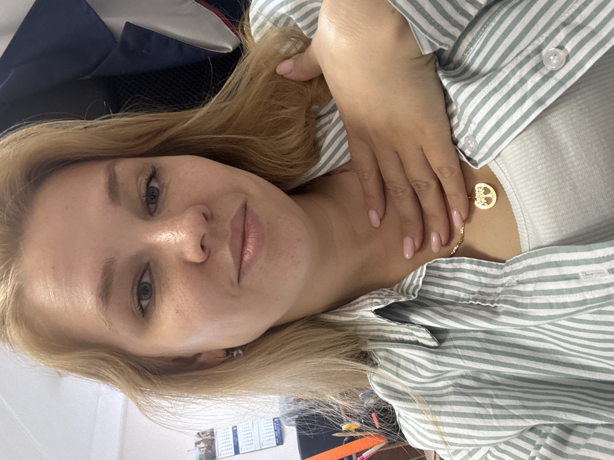 The image is a medium shot selfie of a young Caucasian woman with blonde hair, fair skin, and light green eyes. She is wearing a light green and white striped collared shirt over a white ribbed tank top. A gold necklace with a tree of life pendant is visible around her neck. Her right hand is placed on her chest, with her fingers resting near her collarbone and thumb near her neck. Her fingernails are painted a light pink color.
In the background, a blue and white jacket or fabric is visible to the right. To the left, a wall calendar with several pages is hanging, and a desk or shelf with office supplies such as pens and markers is partially visible. The environment appears to be an indoor setting, possibly an office or a room with office-like furnishings. There are no discernible landmarks or specific features that would indicate a location in the city of Заволжье, Russia. The lighting suggests an indoor environment with artificial or diffused natural light.