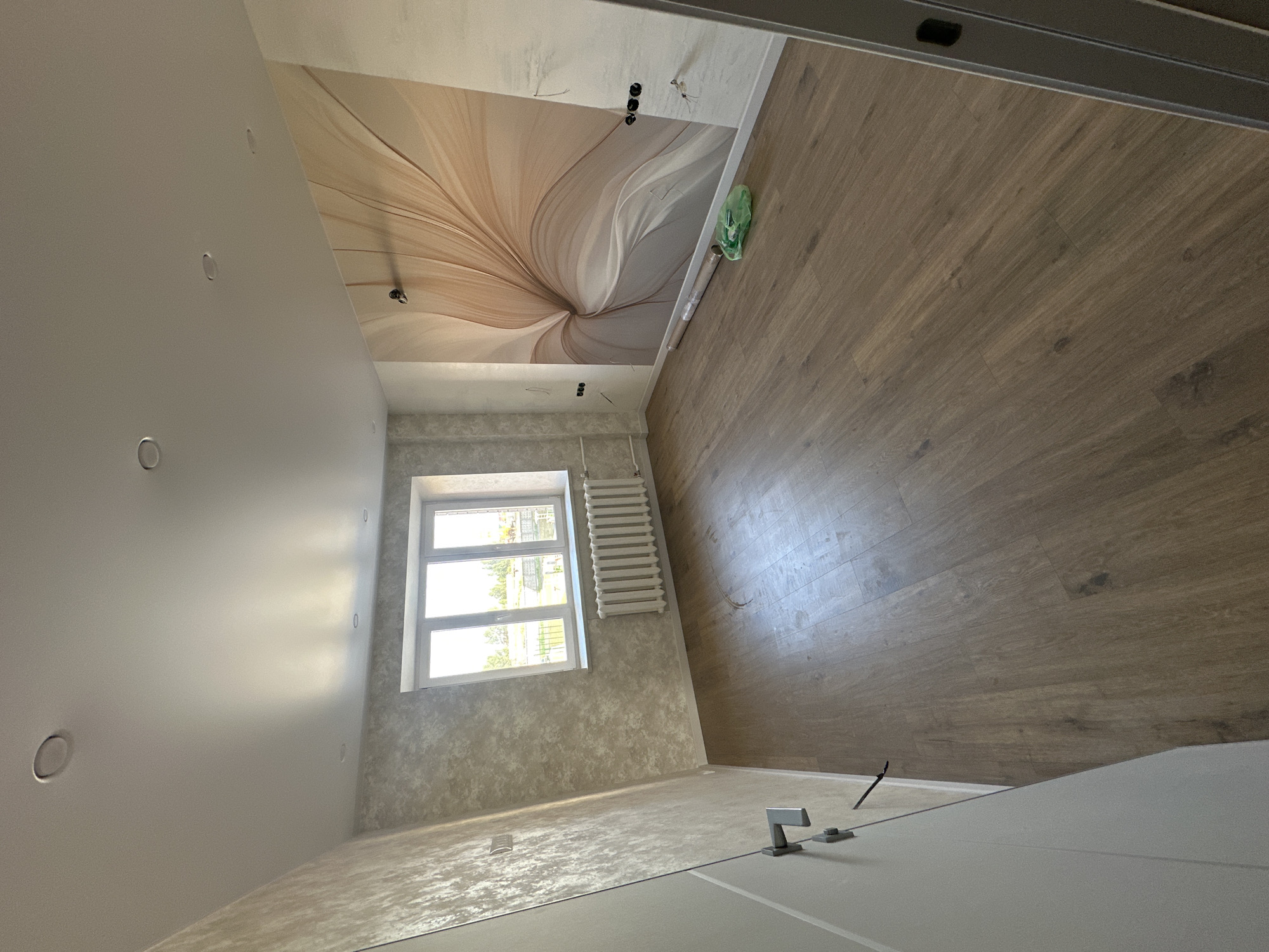 The image shows an empty room under renovation in Novoaltaysk, Russia. The room features a window with a white frame and a white radiator below it. The walls are wallpapered with a textured pattern and one wall has a large floral mural. The floor is made of laminate flooring with a wood grain pattern. There is a white door on the left side of the room with a silver handle. On the right side of the room, near the wall with the mural, there is a rolled-up piece of wallpaper and a green plastic bag. Electrical outlets and wiring are visible on the walls. The ceiling is white and has recessed lighting fixtures. The overall impression is of a room being prepared for occupancy.
