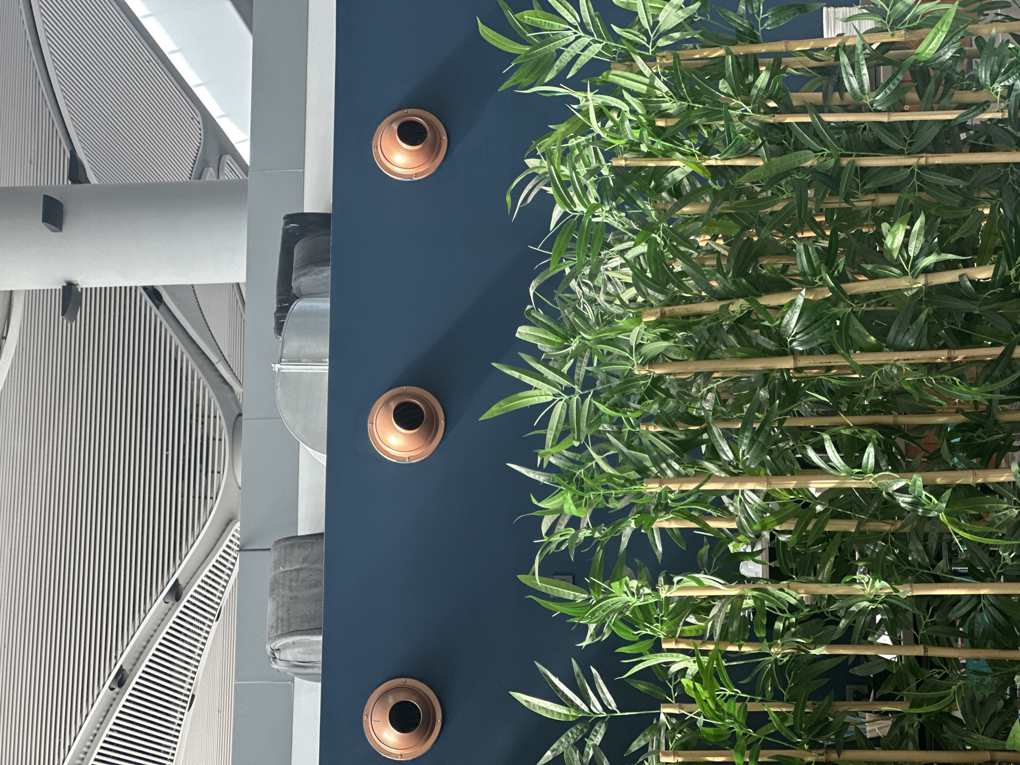 The image shows a close-up of a dark blue wall with three circular copper fixtures mounted on it, spaced evenly apart. Below the wall, a dense arrangement of artificial bamboo stalks and green foliage obscures the lower part of the scene. In the background, above the wall, a portion of a modern ceiling with horizontal slats and curved architectural elements is visible, along with a large white cylindrical pillar. The overall impression is of an indoor space, possibly a public area, with decorative elements and architectural features. The image does not contain any people, actions, or interactions. The location in Arnavutköy, Turkey is not visually indicated within the image itself.