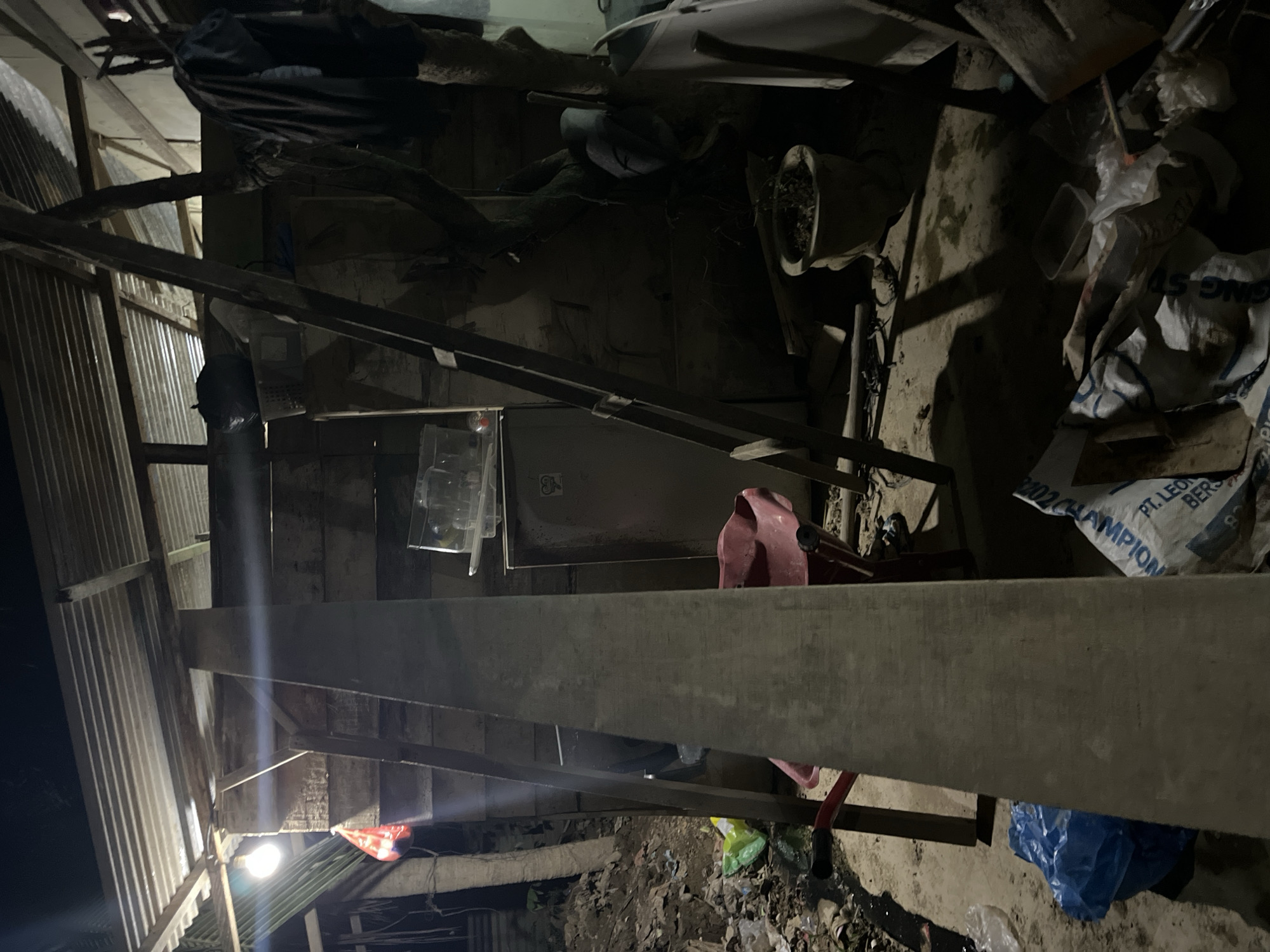 The image depicts a dimly lit, cluttered interior space, likely an outdoor shelter or storage area, located in Benuaraja, Indonesia. A corrugated metal roof is visible overhead, supported by wooden beams. Several objects are scattered around, including a ladder leaning against a wooden wall, a red wheelbarrow, a small refrigerator, and various bags and plastic containers. A bright light bulb illuminates part of the scene. The ground appears to be dirt and debris. The overall impression is one of disarray and a lack of organization.