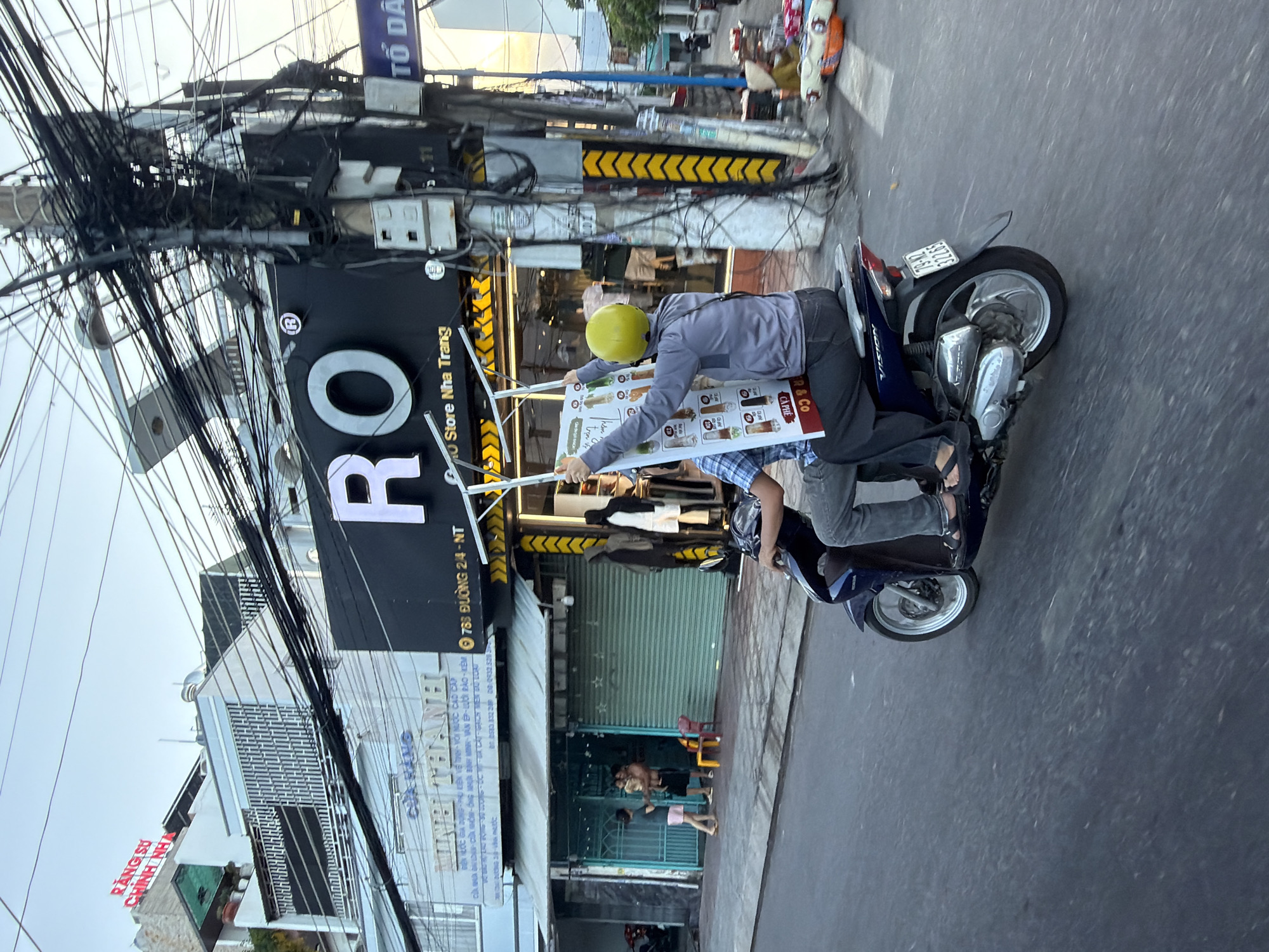The image shows a street scene in Bac Nha Trang Ward, Vietnam. Two people are riding on a blue scooter. The person in the front is wearing a yellow helmet and a grey jacket and is holding up a large advertisement board. The person in the back is wearing a plaid shirt and dark pants. The scooter is driving on a paved road. In the background, there are buildings with signs in Vietnamese, including one that says "RO STORE NHA TRANG". There are many electrical wires hanging above the street, obscuring parts of the buildings. The sky is a pale blue, suggesting it is either early morning or late afternoon.