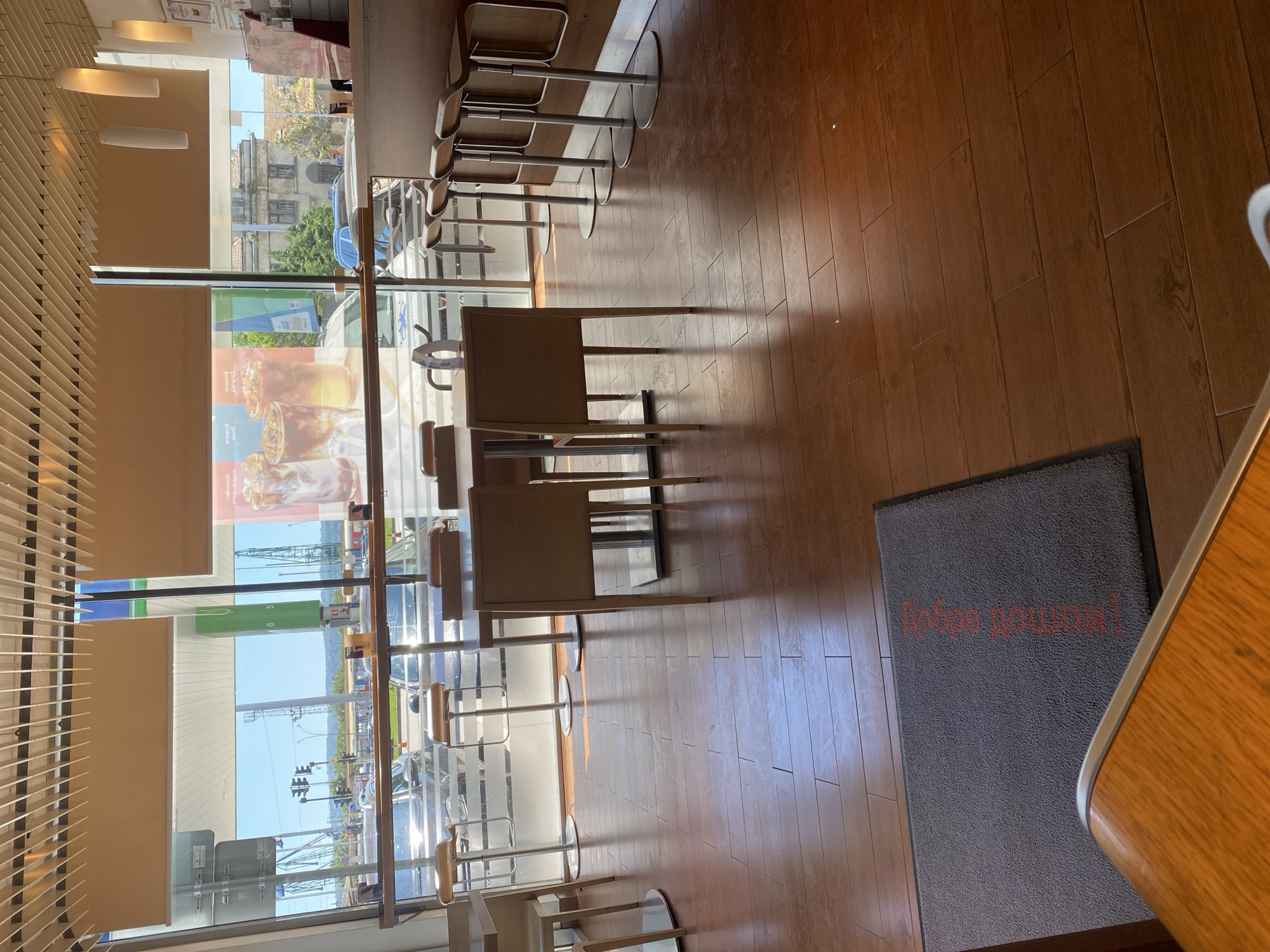 The image shows the interior of a cafe located in Varna, Bulgaria. The cafe features tall glass windows overlooking a street with visible traffic lights, cars, and buildings. Inside, there are high tables and chairs, and a floor mat with the word "Добре дошли!" (Welcome!) written in Bulgarian. Advertisements featuring images of coffee drinks are visible on the windows. There are no people visible in the image.