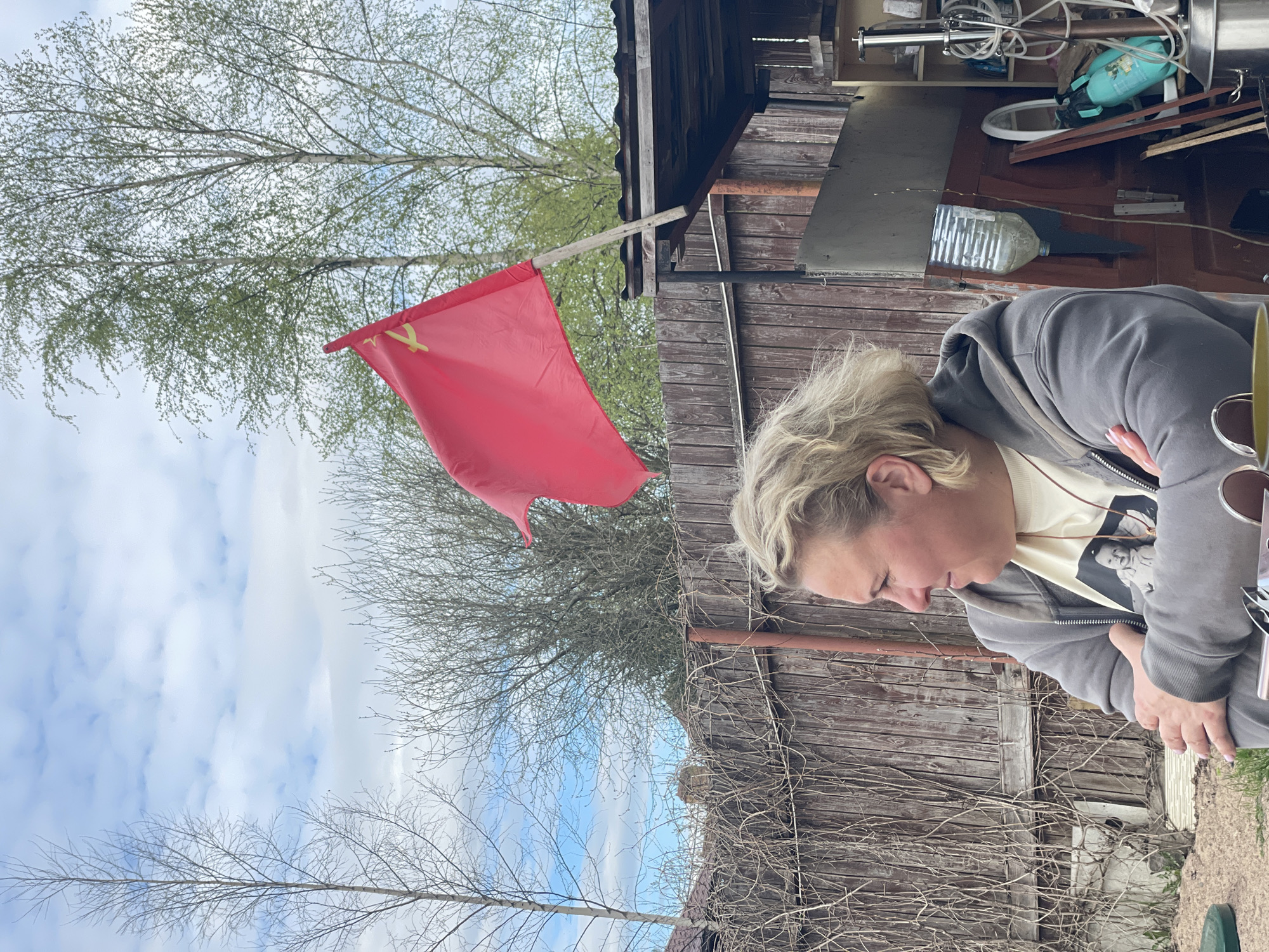 The image depicts a person, presumably female, with short blonde hair and wearing a gray hoodie, appearing to be sitting outside. Behind her is a wooden fence covered with bare vines and a cluttered shed. To the left, a red flag with a yellow hammer and sickle emblem is prominently displayed, attached to a wooden structure. Bare trees and a sky with scattered clouds are visible in the background. The exact location within Sestroretsk, Russia, cannot be determined from the image alone.