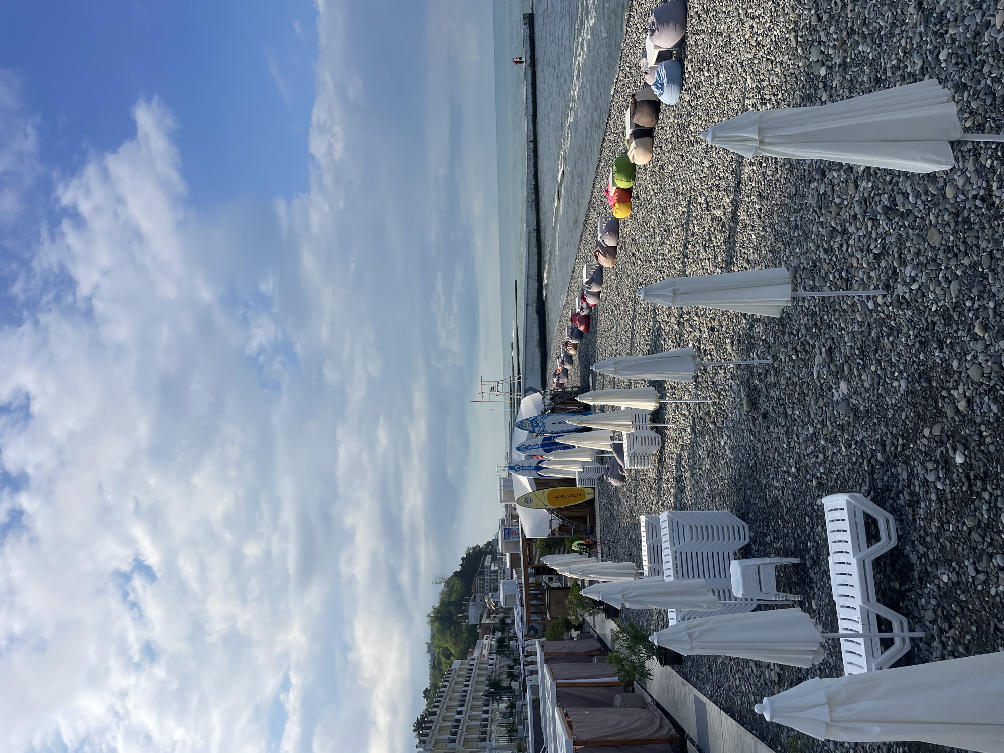 The image depicts a tranquil beach scene on a cloudy day, likely in Sochi, Russia, given the architectural style of the buildings lining the coast and the presence of beach amenities common in the region. The foreground is dominated by a pebbled beach with numerous white, closed umbrellas and stacks of white plastic loungers. To the left, a row of white cabanas with draped fabric walls suggests a designated relaxation area. Further down the beach, a line of colorful beanbag chairs is arranged along the waterfront, some with small tables. In the middle ground, several surfboards and paddleboards are propped upright. The sea is calm with gentle waves lapping at the shore. A concrete pier extends into the water, with two figures visible at its end. In the background, a multi-story hotel with a white facade and balconies overlooks the beach, nestled against a green, tree-covered hillside. The sky is a mix of blue patches and large, white, scattered clouds. The overall impression is one of an organized, yet unoccupied, beach resort, possibly at the beginning or end of the tourist season.