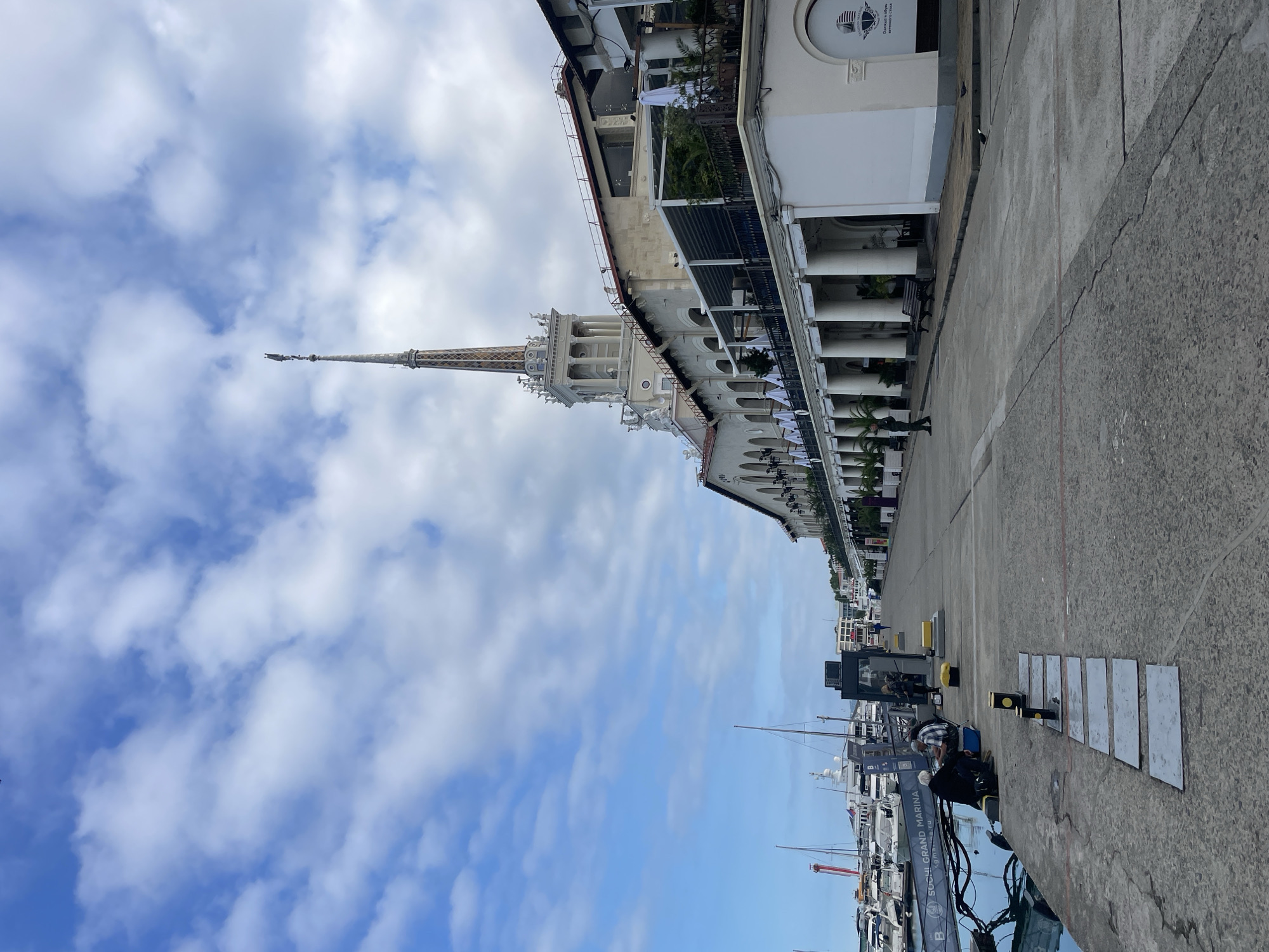 The image shows the Sochi Sea Port in Sochi, Russia. A grand white building with a tall, ornate tower and a spire dominates the right side of the frame. The building features a colonnade with arches along its lower level, some of which are shaded by white awnings. Potted palm trees and lush greenery are visible on balconies and terraces.
To the left of the building, a concrete promenade stretches along the waterfront. Several boats and yachts are docked in the marina, with masts and rigging visible. Some people are seen sitting on the promenade near the water's edge. The sky above is a mix of blue with scattered white clouds. The overall scene depicts a well-maintained port area with a blend of impressive architecture and maritime activity.