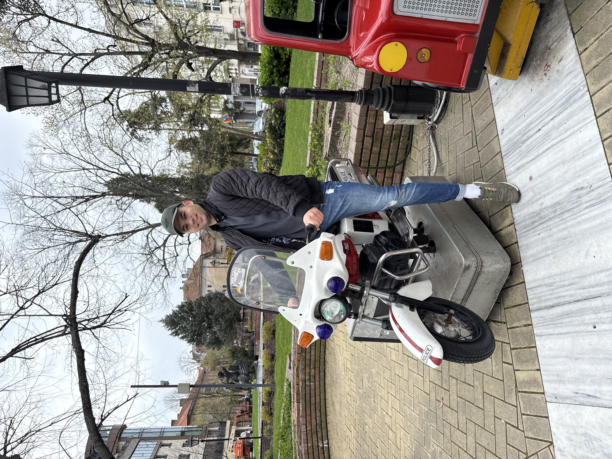 A man sits on a coin-operated children's ride shaped like a police motorcycle. The ride is located on a paved area in a park, next to a red coin-operated ride shaped like a truck or fire engine. A black street lamp stands near the rides. The man is wearing a black jacket, blue jeans, a green hat, and gray sneakers with white socks. He is smiling and looking at the camera. The background includes trees, buildings, and a cloudy sky. This is in the city of Haskovo, Bulgaria.