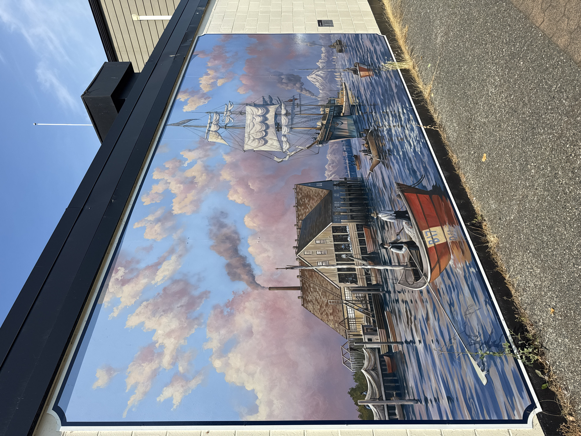 The image is a mural on the side of a building, depicting a historical waterfront scene. The mural features a large sailing ship with full sails on the right side, sailing in a blue body of water. In the background, there are mountains and several smaller sailboats. To the left of the sailing ship, there is a wooden pier with several buildings, including a large structure with a smokestack emitting dark smoke. Several smaller boats are docked or being rowed in the water. Two figures in old-fashioned clothing are visible in a red rowboat in the foreground, to the right of center. The sky in the mural is painted with soft pink and white clouds against a blue backdrop. The building's exterior, to which the mural is attached, has a dark horizontal overhang and a light-colored brick wall on the right. The ground in front of the mural is paved with asphalt and has some dry grass and weeds growing along the edge. This mural is located in Richmond, Canada, specifically on the exterior wall of the Richmond Arts Centre.