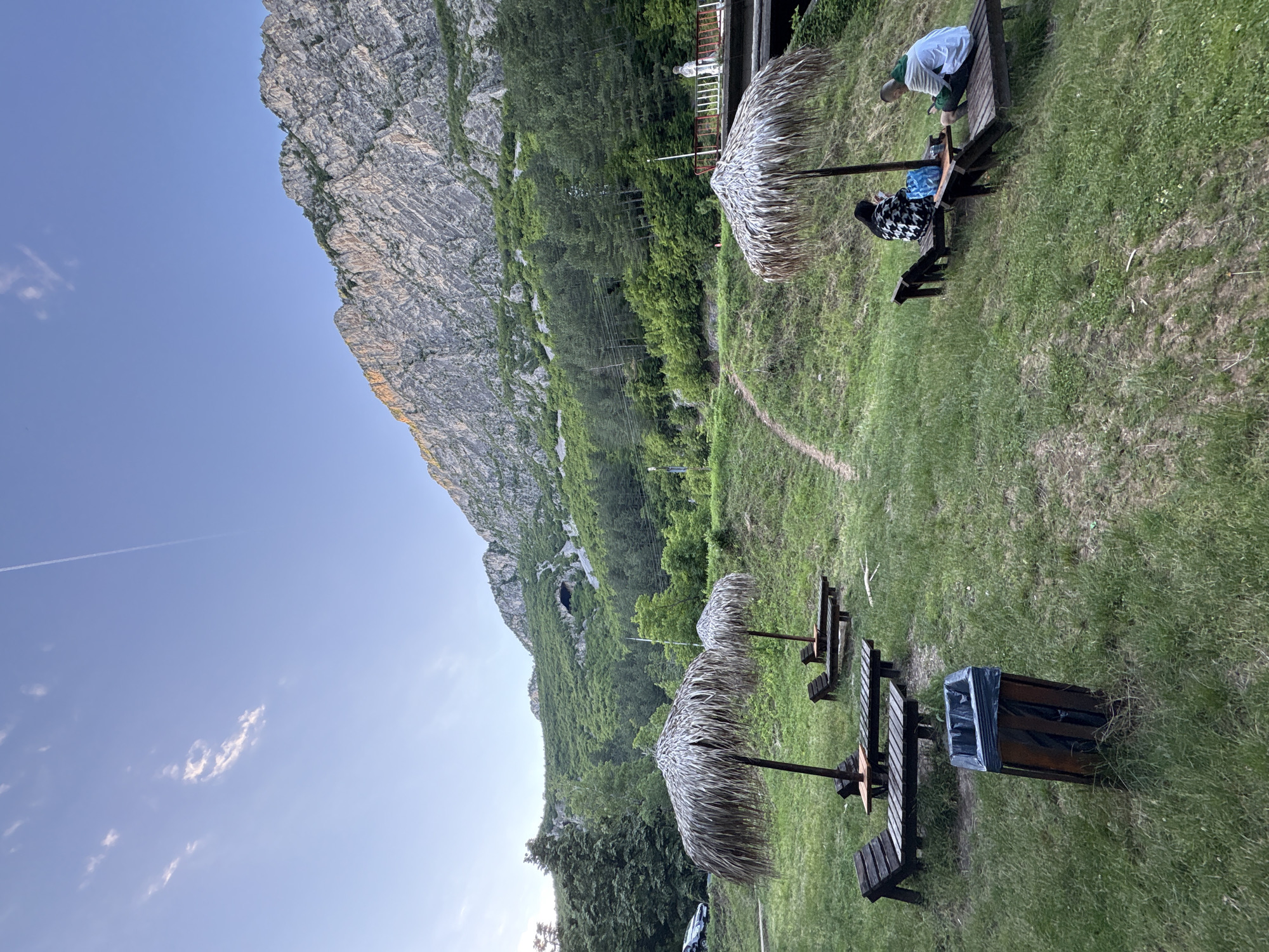 This image depicts an outdoor seating area situated on a grassy slope at the base of a steep, rocky mountain. Several wooden sun loungers are arranged on the grass, some equipped with thatched umbrellas for shade. Two individuals are seated on one of the sun loungers, facing each other and appearing to be engaged with their mobile phones. One person is wearing a black and white patterned jacket, and the other is in a green and white hooded sweatshirt. A small wooden table sits between them, holding a blue bag. A trash can with a black liner is positioned on the grass in the foreground.
The environment is characterized by lush green grass and dense foliage covering the lower slopes of the mountain. The mountain itself is imposing and rocky, with visible striations and textures. A pathway is visible winding up the grassy slope behind the sun loungers. In the background, trees dominate the middle ground, and a large cave opening is noticeable on the mountainside. A road with a parked car is visible to the far left, and a bridge structure with a white railing and a statue is present on the right side of the image. The sky is a clear, pale blue with a few wispy clouds and a faint contrail from an aircraft. The lighting suggests it is either early morning or late afternoon, with sunlight illuminating the upper portions of the mountain.
The scene is located in the vicinity of Zgorigrad, Bulgaria, known for its mountainous terrain and natural beauty. The arrangement of sun loungers and umbrellas suggests a recreational or resting area, possibly a viewpoint or a stop along a hiking trail, taking advantage of the scenic mountain backdrop.