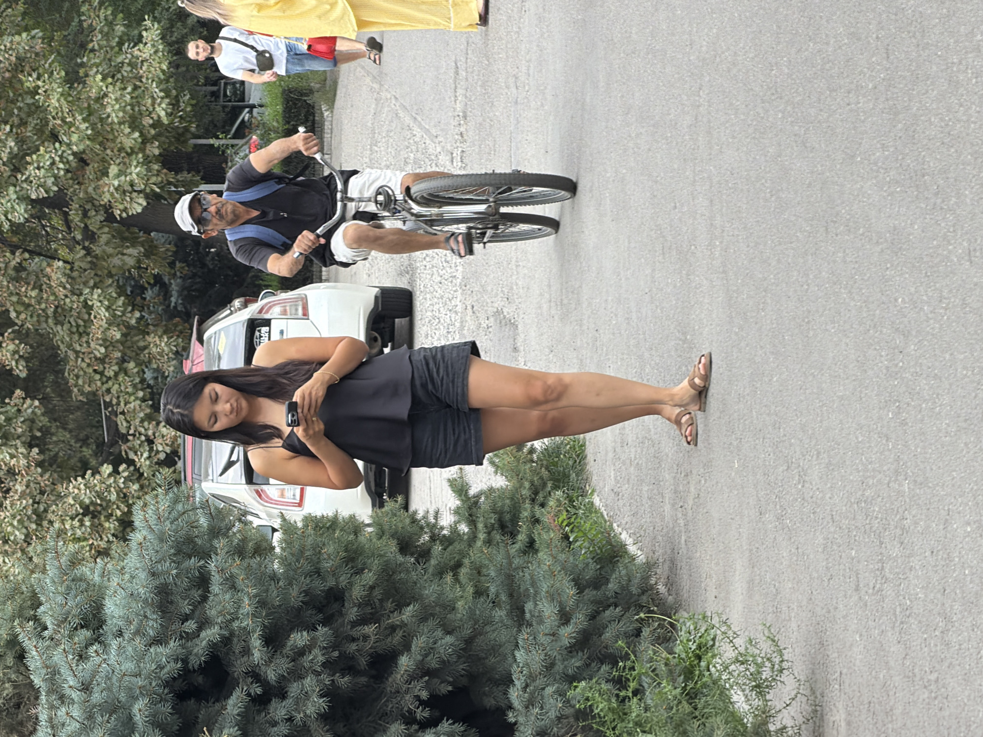 In an outdoor urban setting, a woman wearing a black tank top and black shorts stands on a paved walkway, looking at her phone. She is positioned to the left of a man riding a bicycle towards the right of the frame. The man is wearing a black t-shirt, white shorts, a grey baseball cap, and sunglasses, with a blue backpack. Behind them, a white car is parked. Further in the background, other people are visible, including a man in a white t-shirt and blue shorts walking towards the right, and a person in a yellow outfit. Lush green trees and foliage border the walkway and surround the parked car, suggesting a park or a tree-lined street. The ground is paved with asphalt.