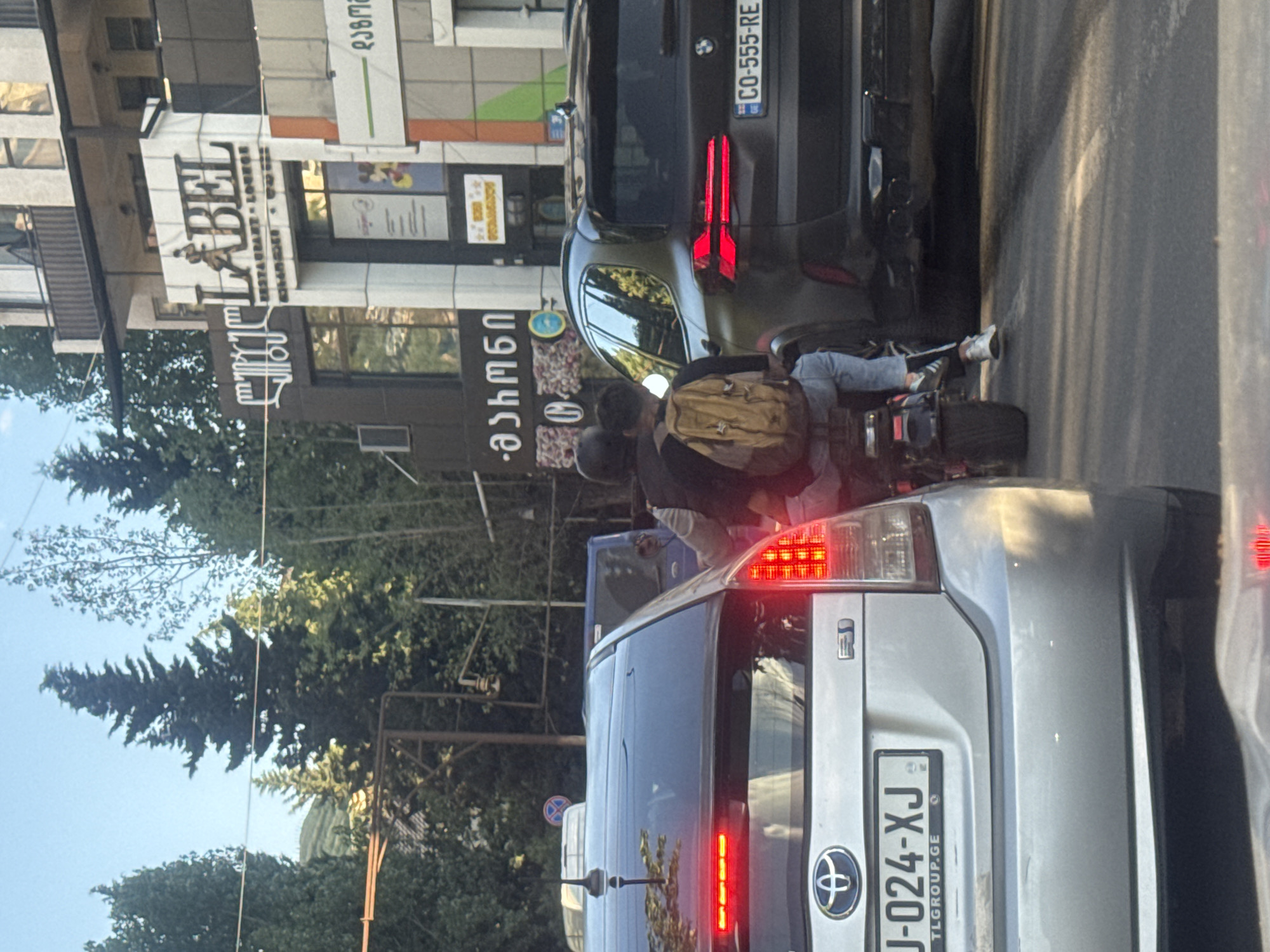 The image depicts a street scene in Tbilisi, Georgia, where traffic is stalled. In the foreground, the rear of a silver Toyota RAV4 is visible, displaying the license plate "J-024-XJ TLGROUP.GE". To its left, a blue vehicle is partially visible. Behind the Toyota, a black BMW SUV is stopped, with its license plate reading "CO-555-RE". A motorcycle with two occupants is positioned between the BMW and the Toyota. The passenger on the motorcycle, wearing a backpack, is leaning back.
On the left side of the image, the facade of a building features signage. One prominent sign reads "LABEL RESTAURANT-LOUNGE", with a stylized depiction of a person in motion. Another sign, in a script that appears to be Georgian, reads "-ashmen". Further in the background, trees and some structures suggest an urban environment. The lighting indicates it is daytime, likely afternoon, with the sun casting shadows. The overall scene captures a moment of traffic congestion on a city street.