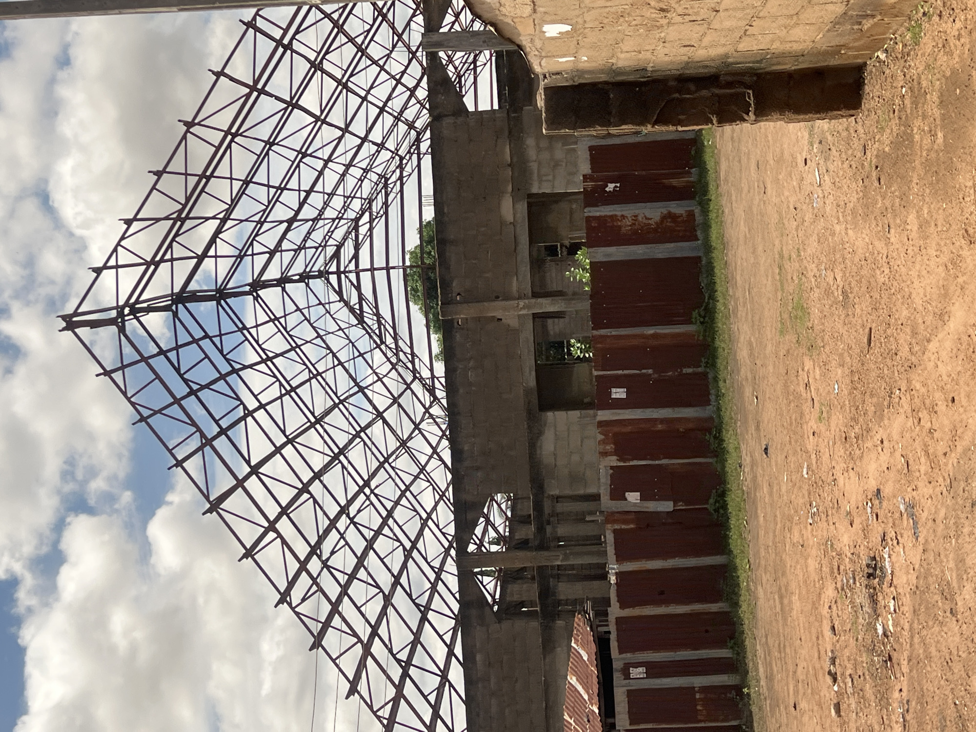 This outdoor image depicts an unfinished building project situated in Jalingo, Nigeria. The upper portion of the frame features a bright blue sky with numerous scattered white and light grey clouds. Dominating the upper-center and spanning across the structure is a large, exposed metal roof truss system, constructed from dark reddish-brown metal beams forming a complex network of triangular segments, typical of a wide-span roof.
Below the roof trusses, the main structure is evident as a multi-level building under construction, primarily built with grey concrete blocks. Visible features include incomplete walls, open window and door apertures, and exposed concrete support columns and beams. On the far left, a small, lower structure with a rusty corrugated metal roof is partially visible.
In the mid-ground, a fence composed of vertically oriented, rusted corrugated metal sheets runs across the scene, appearing to delineate the construction site. Several small white papers are affixed to the surface of this fence. Sparse green vegetation is visible both behind the fence and within the incomplete building structure.
The foreground consists of reddish-brown dirt ground, showing signs of light debris scatter, including small pieces of plastic and paper. On the far right, a partial view of a wall constructed from reddish-brown bricks or blocks is visible, bordering the scene vertically. No individuals, specific actions, or interactions are present in the image. The scene conveys a state of ongoing construction in a daylight setting.