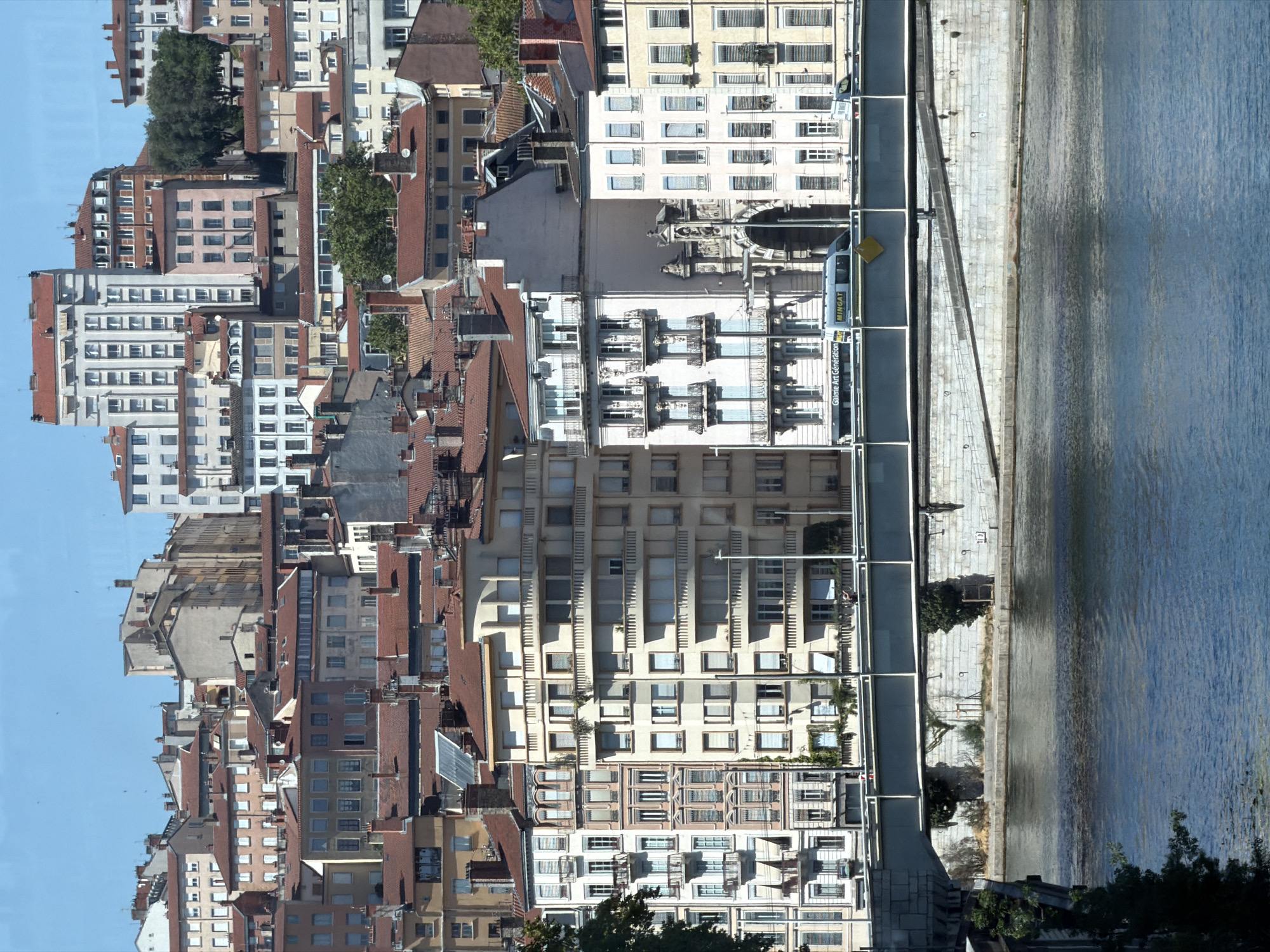 The image shows a panoramic view of the city of Lyon, France, with a focus on its urban architecture and the Saône River. The scene is dominated by a dense cluster of buildings, characterized by their red tile roofs and a variety of architectural styles, ranging from classical to more modern. Many of the buildings are multi-story, with numerous windows and balconies. The foreground is occupied by the Saône River, its blue water reflecting the sky and the surrounding structures. A bridge spans the river, with vehicles, including a van with "MINGAT" branding, visible on it. The bridge's structure is robust, with metal railings and concrete supports. The riverbank is lined with stone walls and some vegetation. The overall impression is of a vibrant, historic city with a strong connection to its waterways.