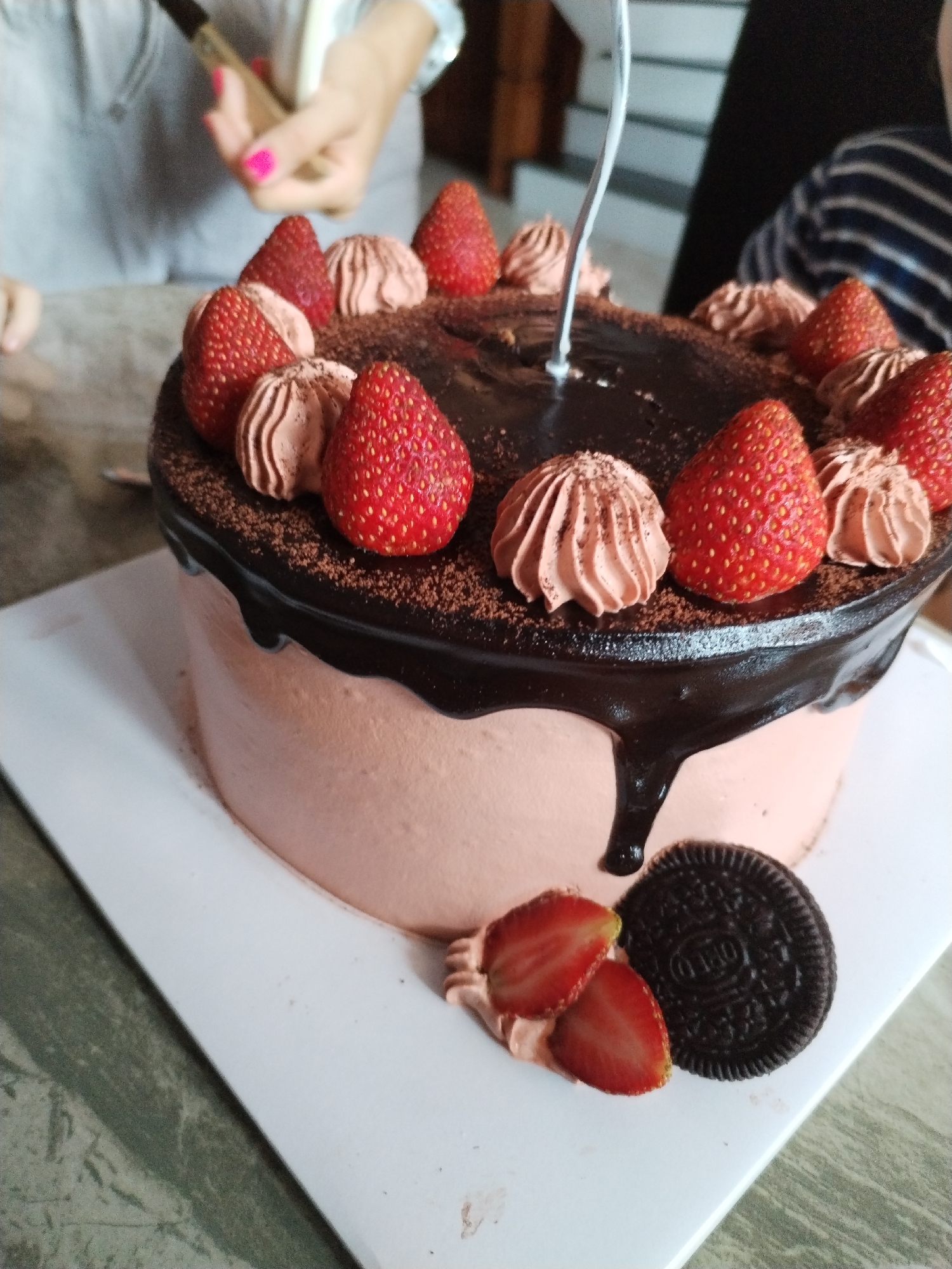 The image depicts a decorated cake. It is a round cake with pink frosting and a chocolate ganache drip. The top is garnished with fresh strawberries and piped chocolate cream swirls. A single Oreo cookie and sliced strawberries sit at the base of the cake. A silver candle is inserted in the center. The image was taken indoors, showing partially visible people on either side of the cake. The location is Bac Nha Trang Ward, Vietnam.