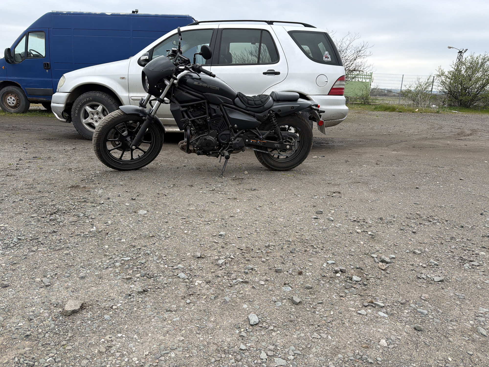 The scene depicts a black Eliminator motorcycle parked on a gravel lot. Behind the motorcycle are a blue van and a white SUV. In the background, there is a partial view of a green fence, some shrubbery, and an electrical pole. The sky is overcast. The location is likely in Burgas, Bulgaria, based on prior information, but there are no obvious landmarks visible in the image to confirm this definitively. No people are visible in the image.