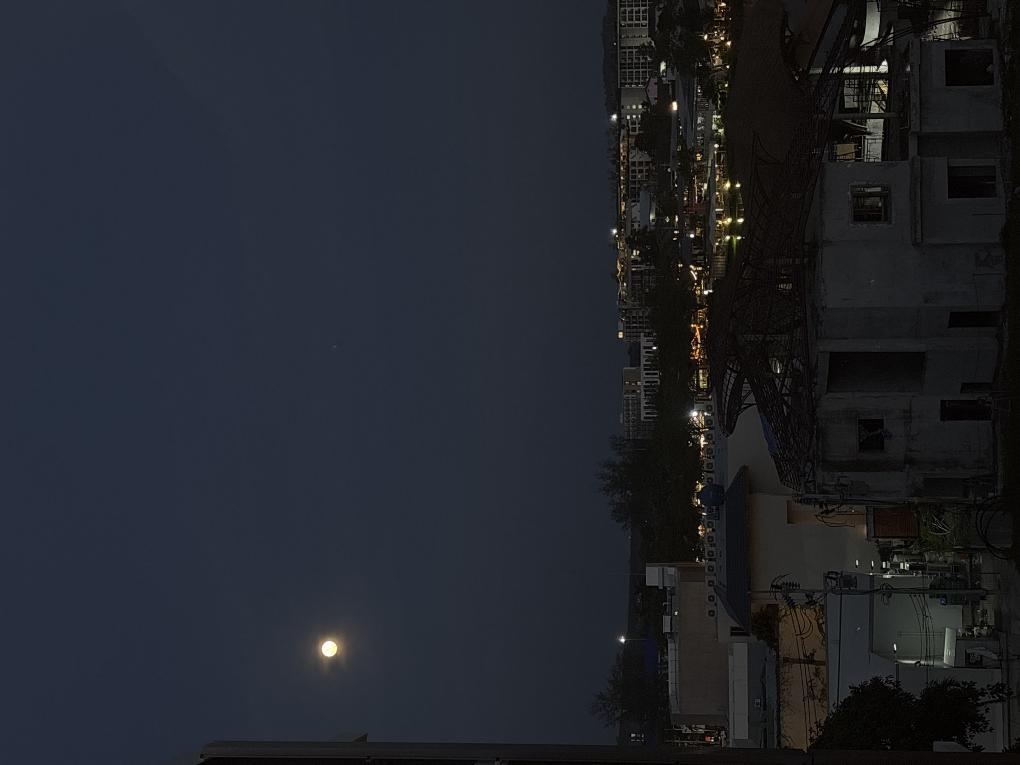 The image shows a night view of Choeng Thale, Thailand. In the lower left corner, the moon is visible in the dark sky. Below it, the dark silhouette of a rooftop is visible. To the right of the sky, there is a blurred view of a city street lined with buildings and street lights. Further to the right, there is an under-construction building. Various other buildings, trees, and infrastructure elements are visible in the foreground. The image appears to be taken from an elevated position, looking towards the city.