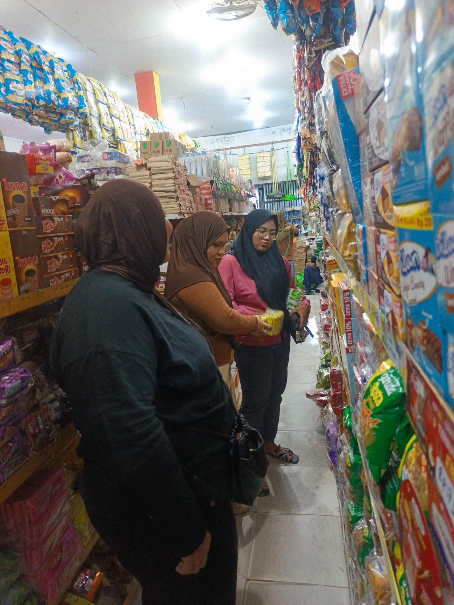 In the city of Ilotidea, Indonesia, a scene unfolds within a brightly lit convenience store. The narrow aisle is lined with shelves overflowing with merchandise, primarily packaged snacks and drinks. Three women are visible. In the foreground, a woman wearing a dark hijab and a dark long-sleeved top stands facing away from the camera, her back to the viewer. To her right, a woman in a brown hijab and an orange sweater is looking at a package of yellow snacks. She is holding one of the packages. Next to her, a woman wearing a black hijab, glasses, and a pink long-sleeved top is also examining the yellow snacks, holding a similar package. A man in a dark blue shirt is visible further down the aisle, sitting on the floor. The shelves are stocked with a variety of items, including boxes of what appear to be instant noodles or snacks, bottles of beverages, and numerous colorful snack bags. The ceiling is illuminated by fluorescent lights, and a ventilation fan is mounted on it. The floor is tiled.