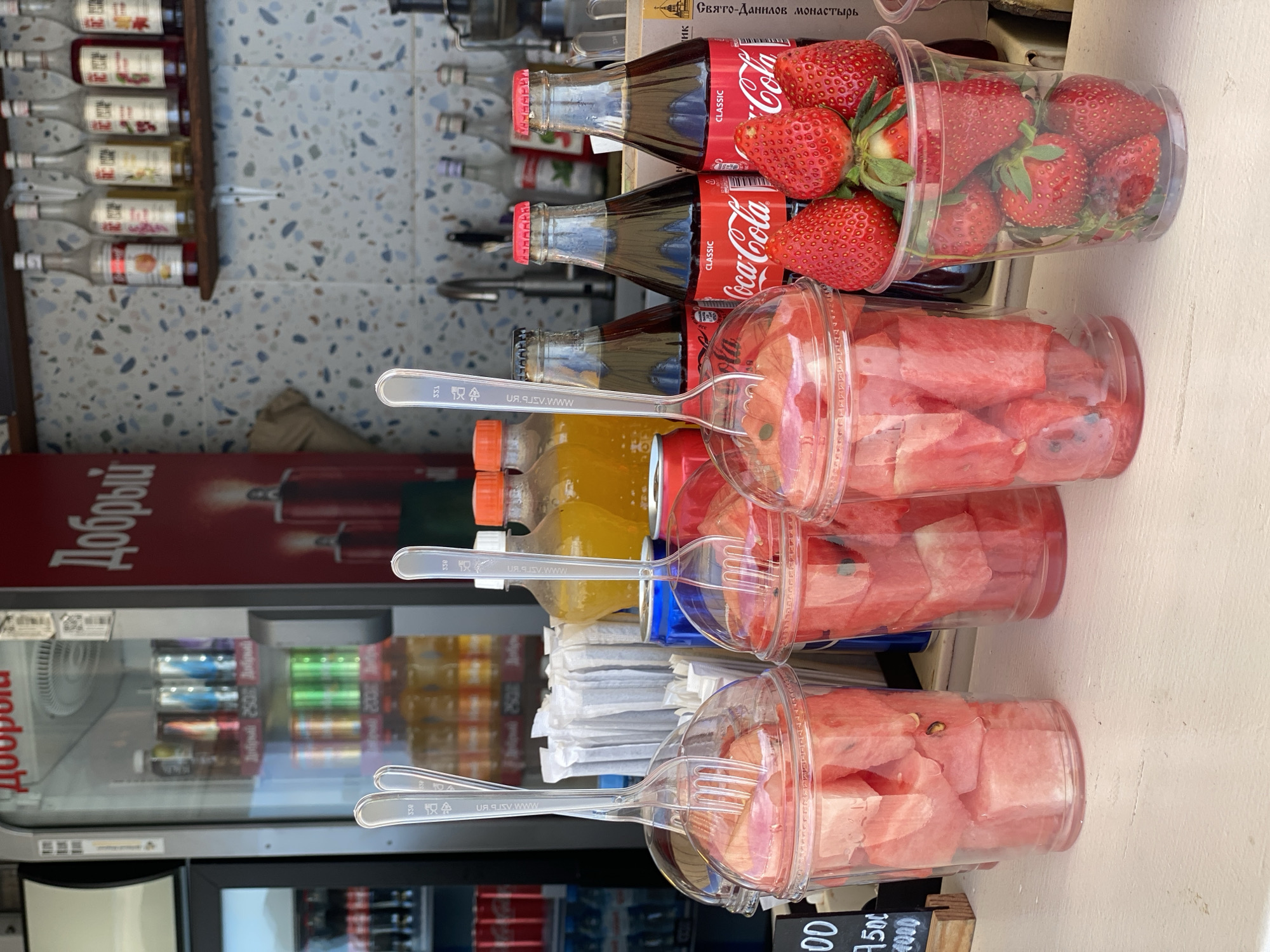 A street stand in Sochi, Russia, features a display of packaged fruits and beverages for sale. Three clear plastic cups filled with cubed watermelon are arranged with disposable plastic forks inserted into each cup. Beside these, two clear plastic cups are filled with fresh strawberries. Three glass bottles of Coca-Cola are positioned alongside the strawberry cups. The stand also has bottles of juice. Behind the display, a beverage cooler is visible, filled with various canned and bottled drinks. To the left, a shelf holds numerous bottles of syrups. "Добрый" is visible on the beverage cooler. The stand is situated outdoors, possibly in a busy tourist area. A sign displaying the prices is also visible.