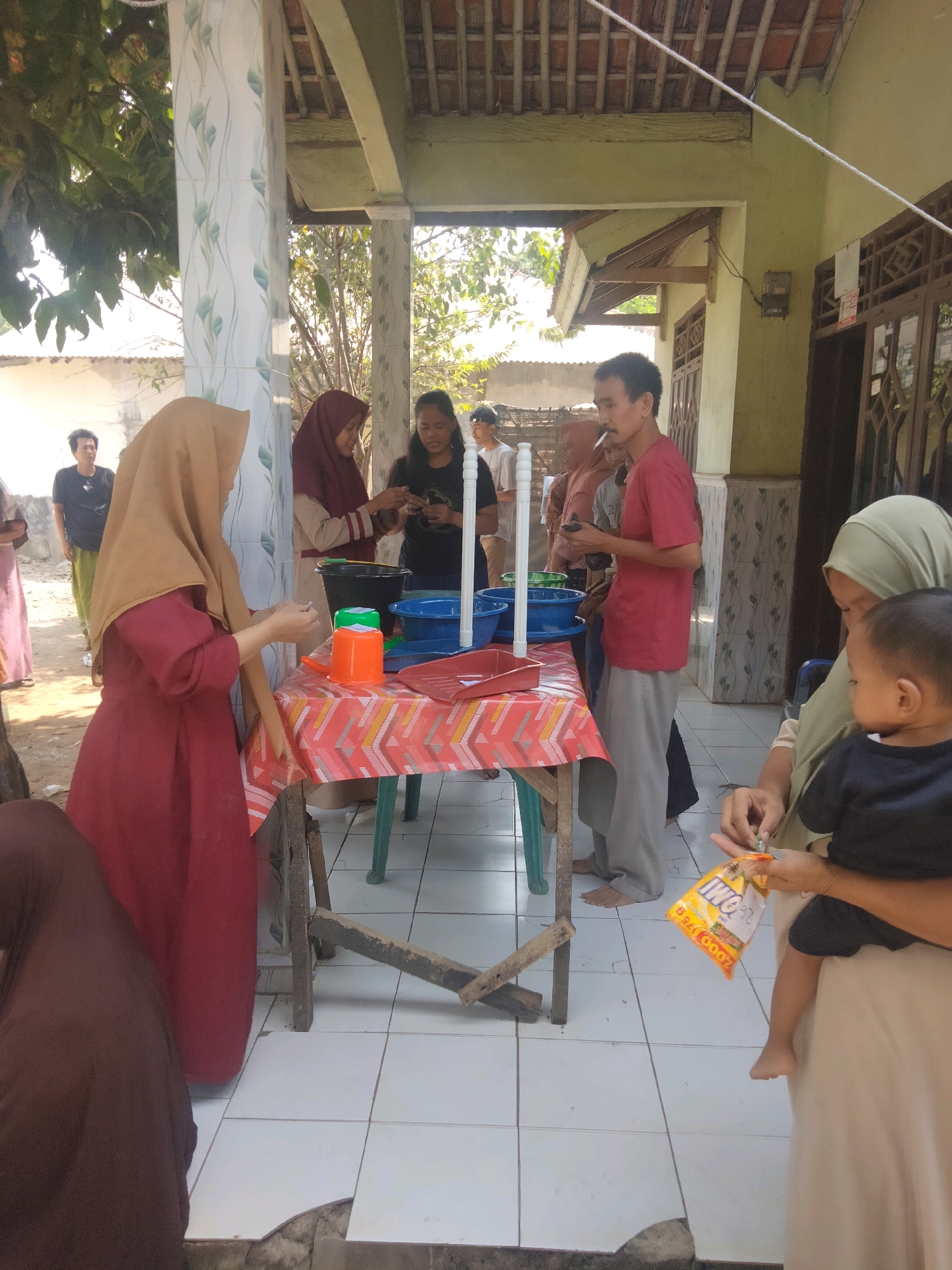 The image depicts a group of people gathered outdoors in what appears to be the city of Suka Sari, Indonesia. A table covered with a colorful, patterned cloth is central to the scene, with various items on it, including plastic basins, cups, and a red tray. Several individuals are interacting around the table, with some appearing to be preparing or distributing something. In the foreground, a woman in a maroon hijab is holding a baby. Behind the table, a man in a red shirt is smoking a cigarette. The background reveals a building with a tiled roof and a white wall, suggesting a residential or community setting. The overall atmosphere seems to be one of an outdoor gathering or event.