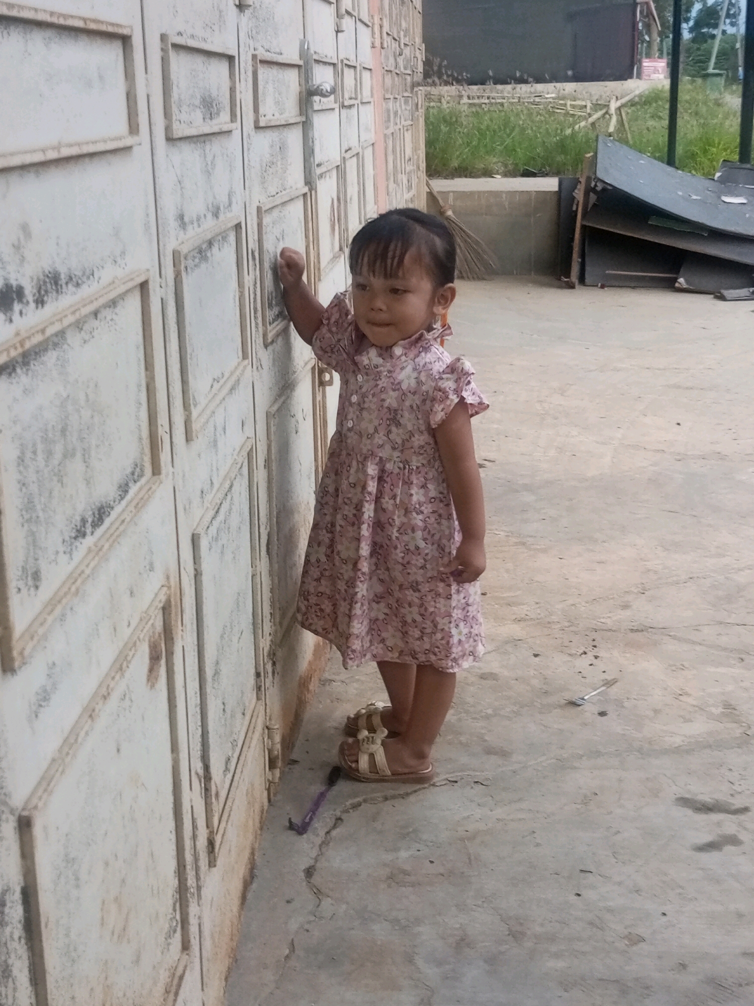 A young girl, wearing a pink floral dress and sandals, stands in front of a white, weathered metal door. She is positioned on a concrete surface. To her right, beyond the door, is an area with overgrown grass and some discarded materials, including what appears to be sheet metal and wood. The scene suggests an outdoor setting in what could be a residential or storage area. The specific location in Bundar, Indonesia, is not determinable from the visual information provided.