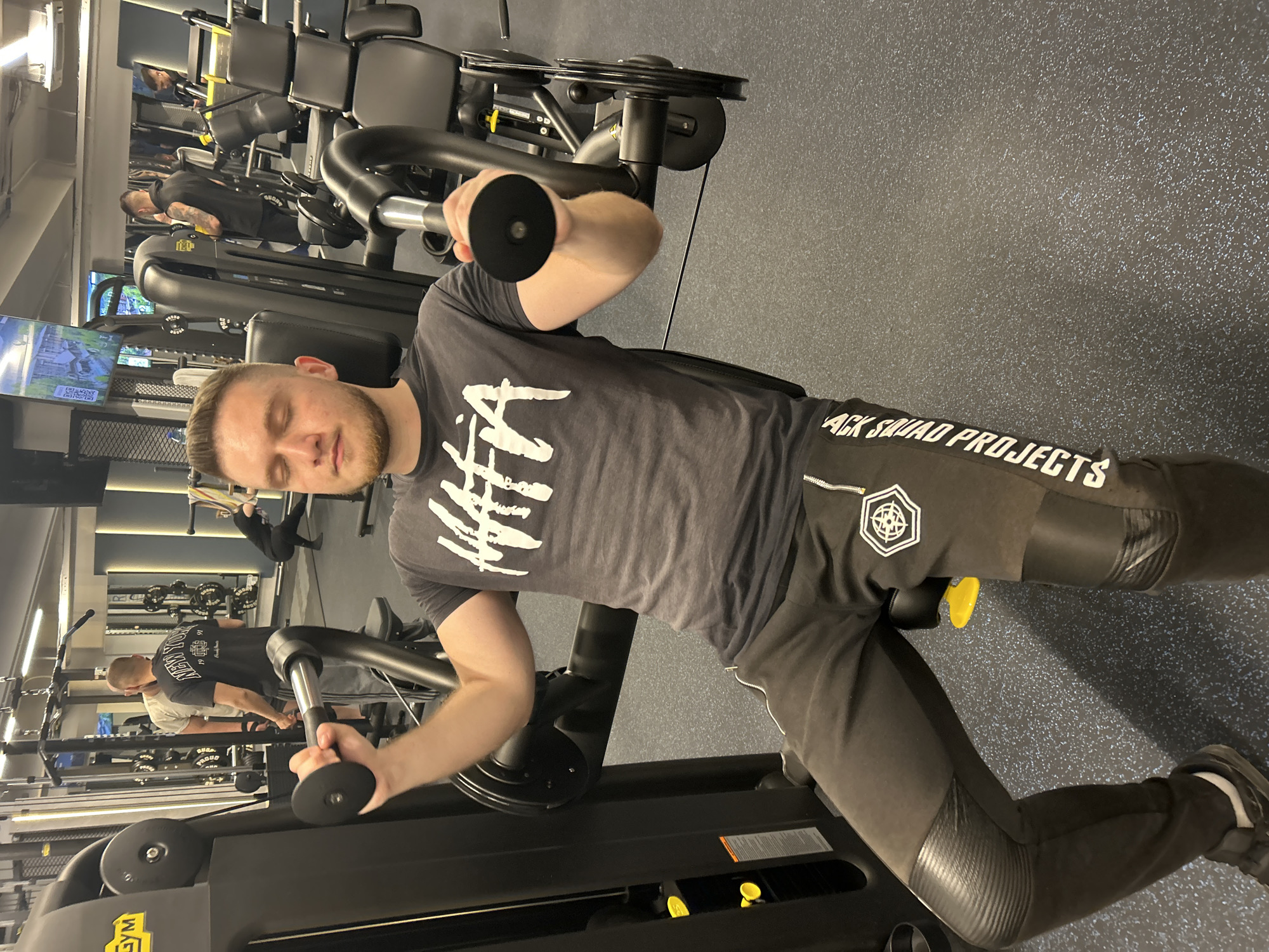 The image shows a man in a gym using a chest fly machine. He is wearing a gray t-shirt with a white design and black pants with a logo and text on the right leg that says "ACK SQUAD PROJECTS". He is sitting on the machine with his eyes closed and is holding the handles. The gym has modern equipment, a gray and blue floor, and mirrors on the wall reflecting the scene. The location is unspecified and has no indication of being in the city of Varna, Bulgaria.