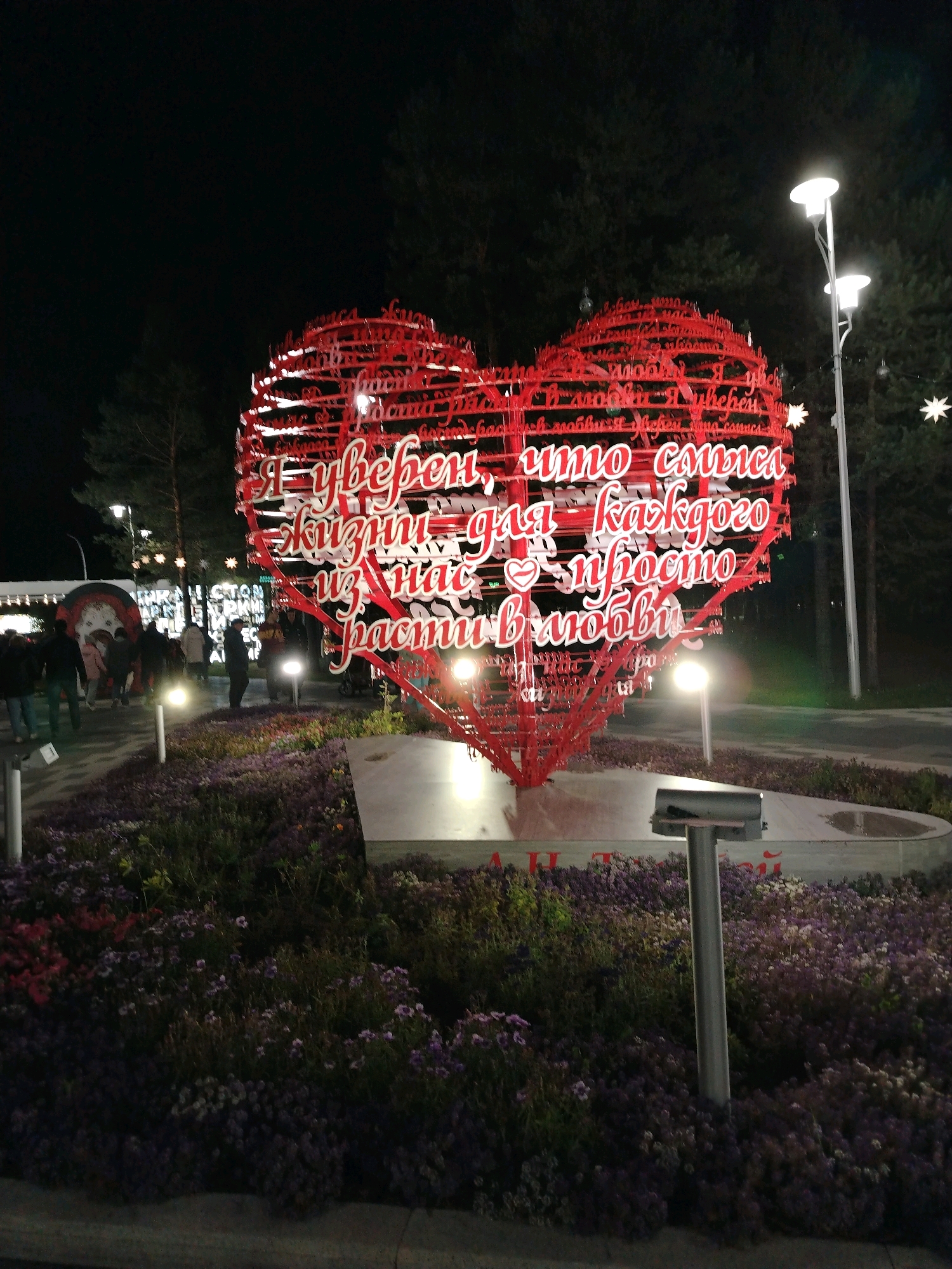A large, red, heart-shaped sculpture with white text is illuminated at night in Muravlenko, Russia. The text on the sculpture, which appears to be a quote in Russian, is comprised of white, three-dimensional letters. The sculpture is situated in a park or public space, with a bed of purple and pink flowers surrounding its base. Several lampposts with bright lights are visible, casting a glow on the scene. In the background, trees can be seen, and a few people are walking in the distance, some appearing to be illuminated by the lights of the sculpture. The overall atmosphere is festive and romantic.