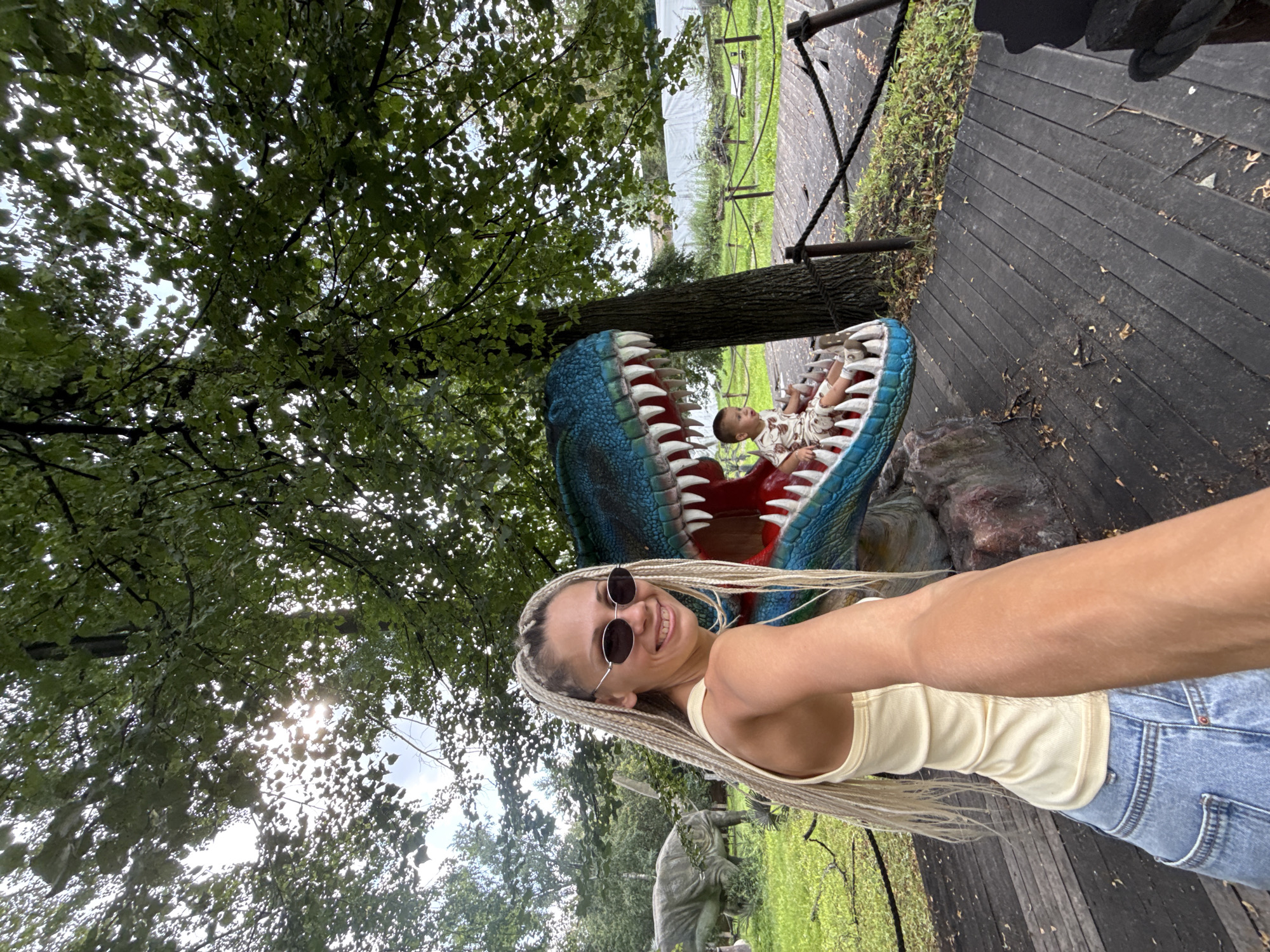 A woman with blonde braided hair and sunglasses smiles at the camera while taking a selfie. She is wearing a yellow tank top and blue jeans. To her right, a large, blue dinosaur mouth with sharp white teeth is visible. A young boy in a patterned outfit is sitting inside the dinosaur's mouth, looking to the left. In the background, a large, grey dinosaur statue stands in a grassy area. The scene appears to be an outdoor exhibit, possibly a dinosaur park, with wooden walkways and trees. The setting is not specified to be in the city of Zhukovo, Russia.