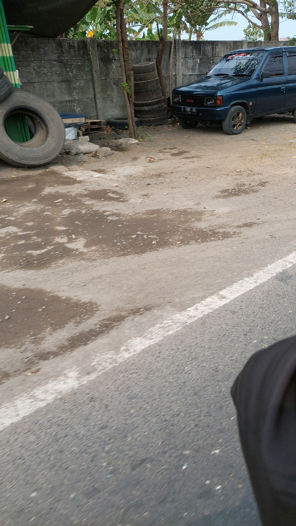 In Sidoarjo, Indonesia, a dark blue SUV is parked on a dirt lot next to a concrete wall. The SUV is facing towards the left of the frame, and its tires are a bronze color. In the foreground, a large tire is lying on its side. A white stripe runs across the asphalt in the lower right portion of the image. Behind the SUV, a stack of tires leans against the wall. The ground is a mix of dirt and puddles, with some scattered debris. The scene appears to be outdoors, possibly in a storage or maintenance area.