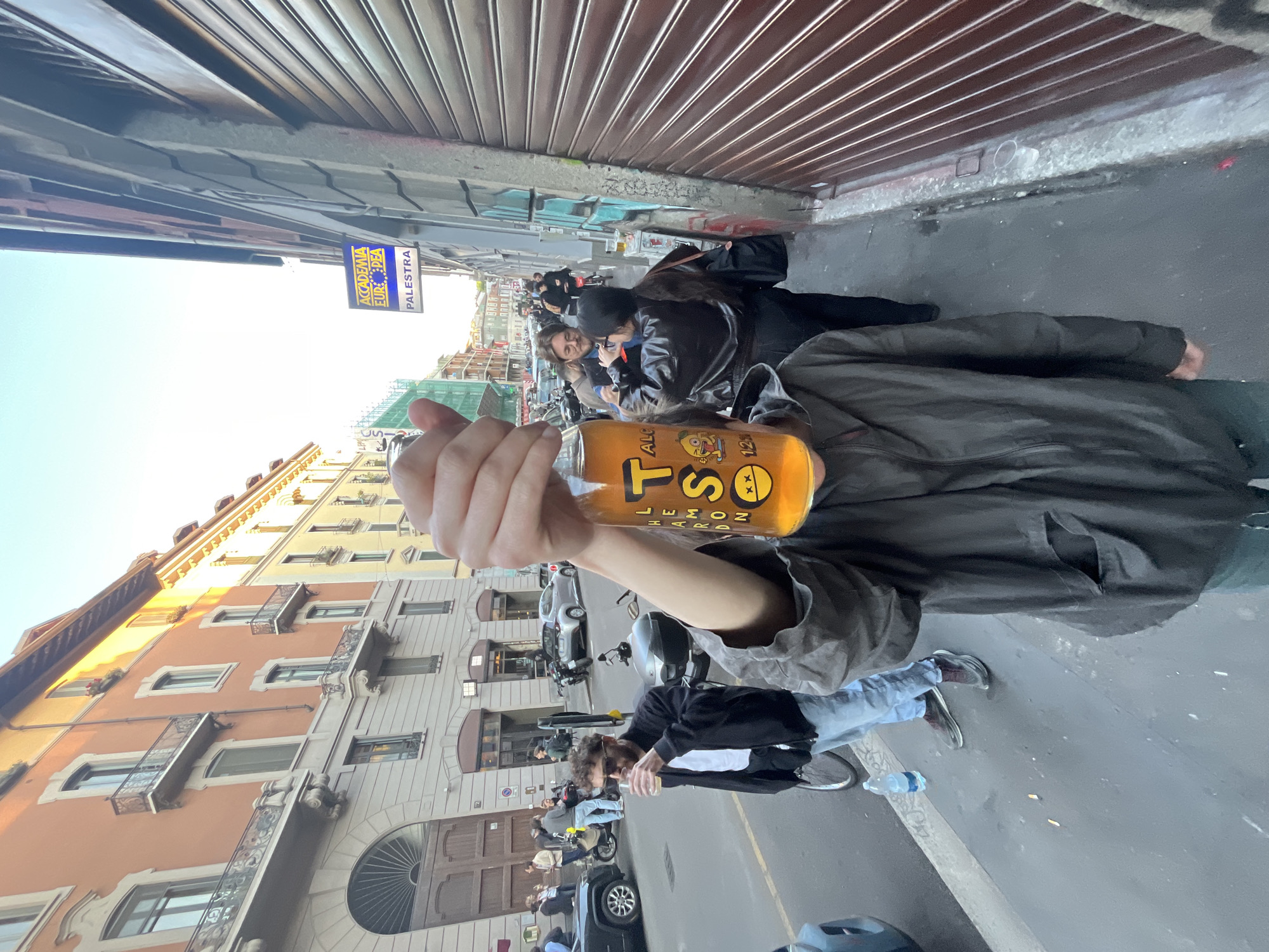 The image, rotated 90 degrees clockwise, presents an urban street scene from a low angle. In the foreground, a person holds an orange-colored bottle with their hand and arm visible. The bottle's label clearly shows "TS," "HARD LEMON," and "ALC 1.2%," along with a cartoon character featuring crossed-out eyes. The person holding the bottle is wearing a dark-colored jacket or coat.
The background reveals a narrow city street lined with buildings. On the left side of the street, a metallic roller shutter, likely belonging to a closed shop, extends along the base of a building. On the right, multi-story buildings with balconies, showcasing traditional European architecture in warm tones of orange, yellow, and grey, recede into the distance. A bright, clear sky is visible above the rooftops.
The street is populated with several people walking and standing, alongside parked motorcycles, scooters, and a few cars. A blue and yellow sign, attached to one of the buildings, is visible, displaying the text "ACCADEMIA EUROPEA" and "PALESTRA," indicating an Italian-speaking location. The scene appears to be taking place during daytime, with clear weather conditions. A discarded plastic water bottle is visible on the ground near the feet of a person further down the street.