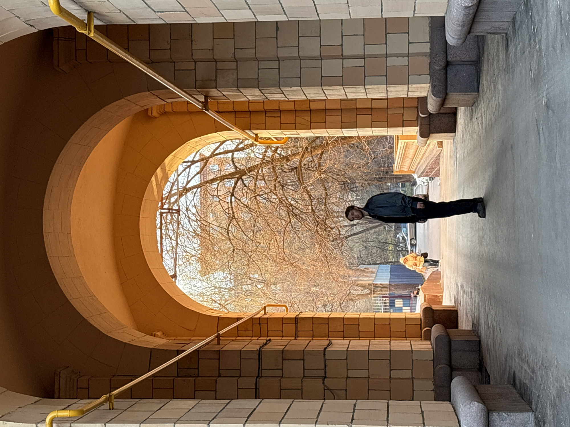 A man stands in a long archway of brick and concrete in Moscow, Russia, on a clear day. The archway has a large, curved opening at the far end through which bare trees are visible. A second person is further down the walkway wearing a yellow jacket. Yellow metal pipes line the top of the archway. The walkway is made of light grey concrete and has decorative stone elements on either side.