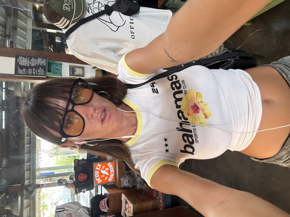 A woman with dark hair, bangs, and yellow-tinted glasses poses close to the camera wearing a white t-shirt with yellow trim, a floral design, and “Bahamas” printed on the front. She has headphones in her ears and silver hoop earrings. A black bag is slung over her shoulder. In the background, there’s merchandise displayed on shelves and what appears to be a mirror reflection. The overall composition is casual and selfie-style.