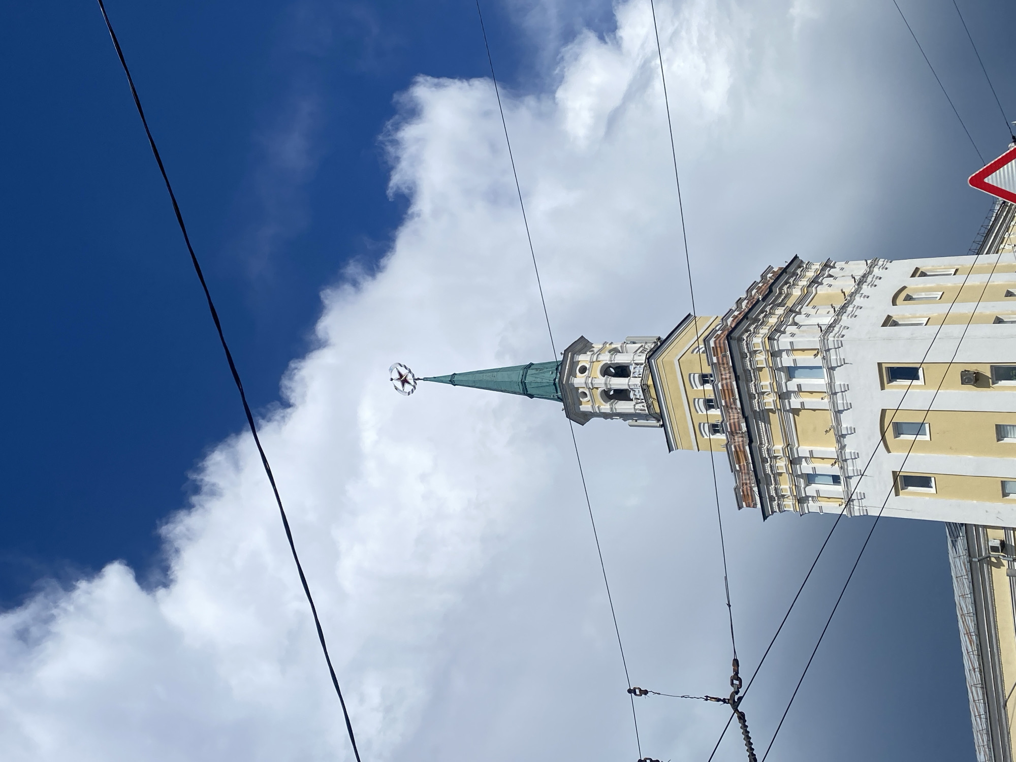 This media file depicts an upward view of a distinctive building with a spire, set against a sky filled with dramatic clouds. The building is predominantly yellow and white, with ornate architectural details and multiple windows. The spire, a striking green color, is topped with a red star. The presence of overhead wires, including some with visible insulators and chains, suggests an urban or semi-urban environment, possibly near a tram line. A red and white triangular traffic sign is partially visible in the upper right corner. The sky is a mix of deep blue and bright white clouds, indicating a bright, possibly windy day. The perspective, looking upwards from a low angle, emphasizes the height of the building and the expanse of the sky. The overall mood is one of grandeur and architectural interest, with the sky adding a dynamic element to the scene. No people are visible in the frame. The location context of Заволжье, Russia, combined with the architectural style and the red star on the spire, hints at a location with historical or Soviet-era significance. There is no discernible text in the image.