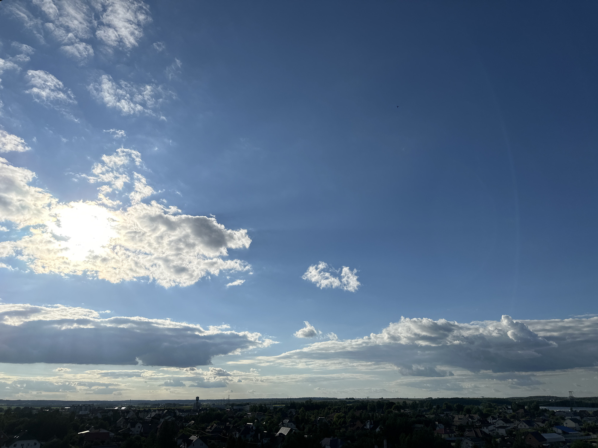 The image captures a wide, expansive view of the sky above a rural area, likely on the outskirts of the city of Шчомысліцкі сельскі Савет, Belarus. The sky is a vibrant blue, dotted with a variety of clouds. On the left side, a bright sun is partially obscured by fluffy, white cumulus clouds, casting rays of light through the atmosphere. Towards the center, a few smaller, scattered clouds drift across the sky, and a tiny, dark object, possibly a bird or an insect, is visible in the upper right quadrant of the image. The horizon is lined with a dense, dark green expanse of trees, below which are clusters of residential buildings, indicating a village or suburban settlement. The rooftops of houses are visible in varying shades, and the overall impression is one of a peaceful, daytime scene in a country setting.