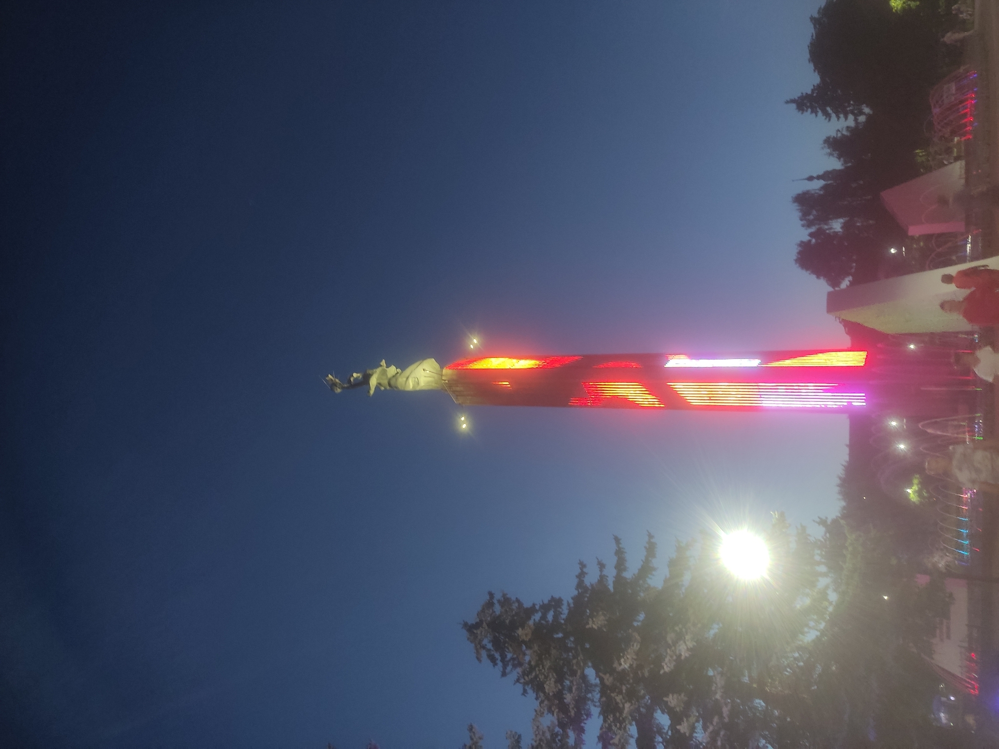 This image depicts the evening sky over the city of Kaluga, Russia, with a prominent monument illuminated by vibrant red and yellow lights. The monument, likely a memorial, features a statue at its apex. A large tree, illuminated by a bright light source that creates a sunburst effect, is visible to the left of the monument. In the foreground, a fountain with colorful lights sprays water, and several individuals are present, though their features are indistinct due to the lighting and distance. The overall scene suggests a public space in Kaluga at dusk or night.