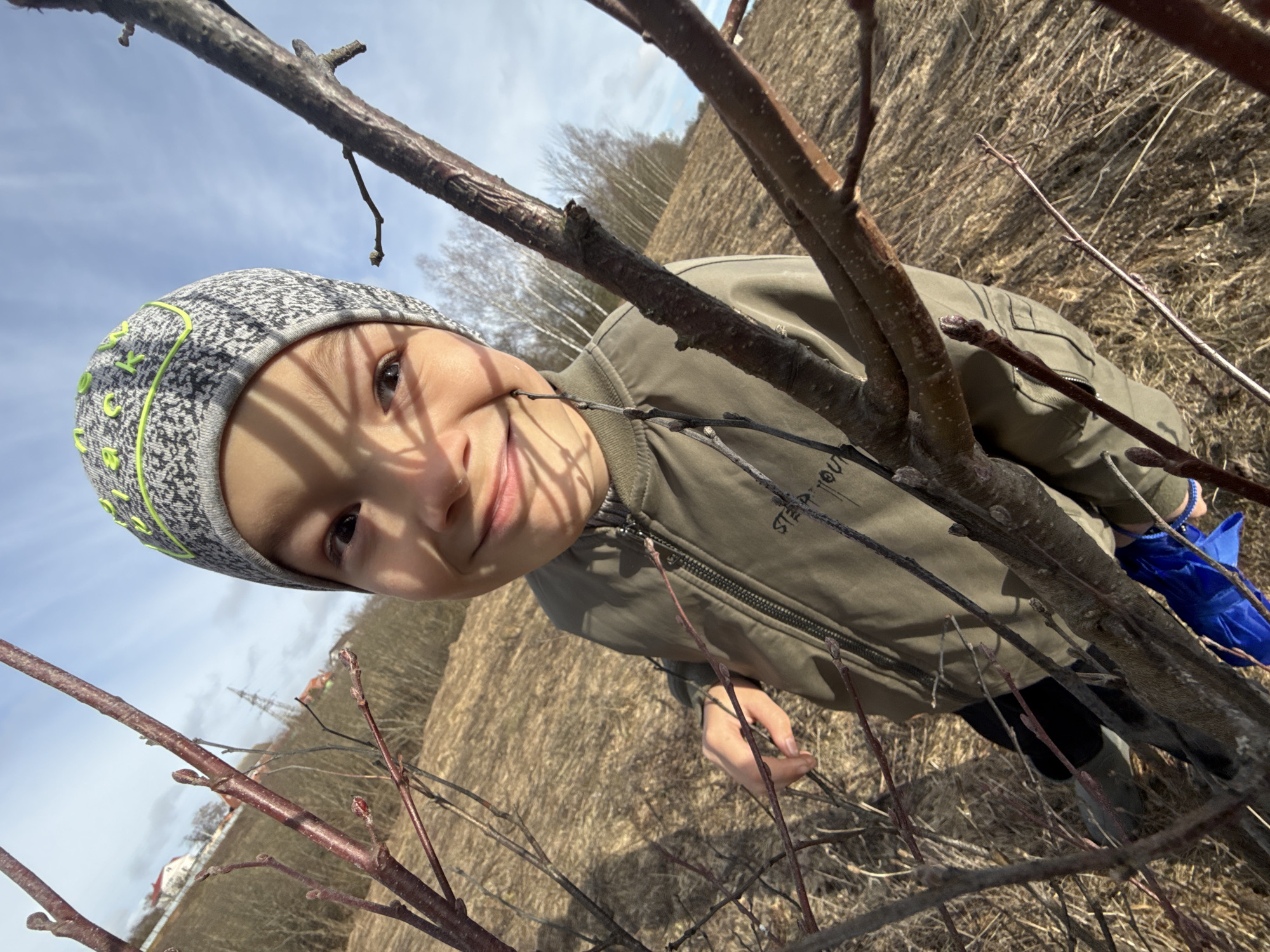 A young child wearing a gray hat with neon green trim and a green jacket is positioned amongst bare branches, presumably in an outdoor setting. The background suggests a field or undeveloped area. The child is smiling slightly at the camera, holding a small branch. The scene gives no clear indication of being Ignatovka, Russia. No landmarks or specific details confirm this location. A blue object is held on the right side, behind the branch.
