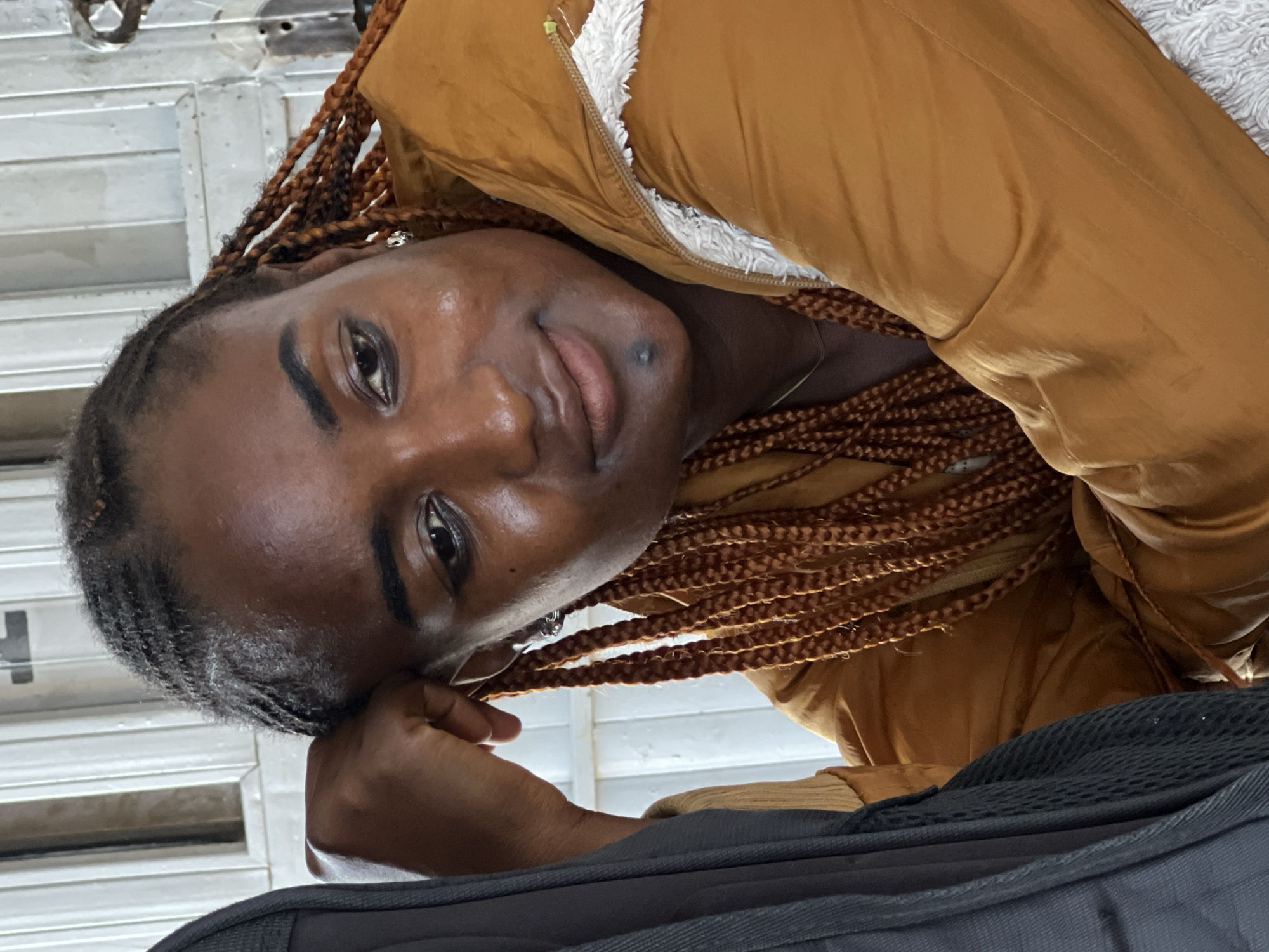 A woman with braided dark hair, resting her head on her hand, is looking down and to the right. She is wearing a rust-colored jacket with a white fluffy lining around the collar. A dark backpack is visible on the left side of the frame, partially obscuring the woman and the background. The background appears to be an indoor setting with white vertical paneling and possibly a window or door frame, suggesting a location in Jos, Nigeria. The lighting is soft and diffused, creating a natural and candid feel to the image. The woman has a small mole on her chin.