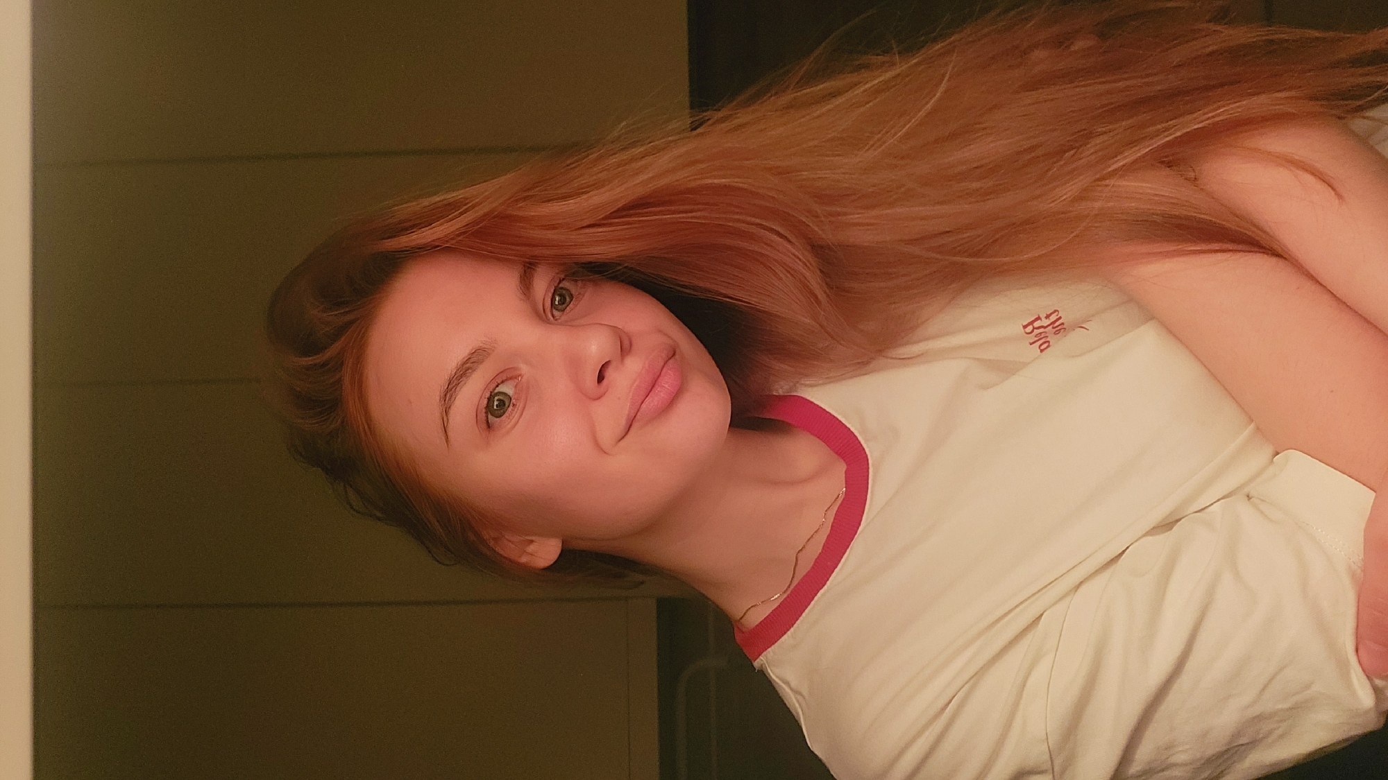 A young woman with long, light red hair is posing for a selfie or casual photograph. She is wearing a white t-shirt with a red neckline and a small red graphic on the chest. Her hair is spread out, occupying a significant portion of the frame. She has fair skin and is looking directly at the camera with a slight smile. A thin gold necklace is visible around her neck. The background is blurry and dimly lit, appearing to be a wall. The image is taken indoors. No details are available to identify the specific location in Chaikovsky, Russia.