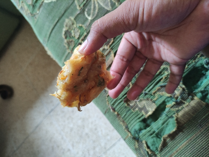 The image shows a hand holding a piece of fried food, possibly a fritter or a pastry. The hand is holding the food up against a green fabric surface, which has a pattern of stripes and is torn in places. The food is crispy and brown, and it appears to have a filling of some kind. The scene is most likely a domestic one, with a sofa or similar furniture in the background. The coordinates provided, -7.828 latitude and 112.701 longitude, place the location in Kecamatan Lawang, Indonesia. There is no additional information available to provide further context about the scene.