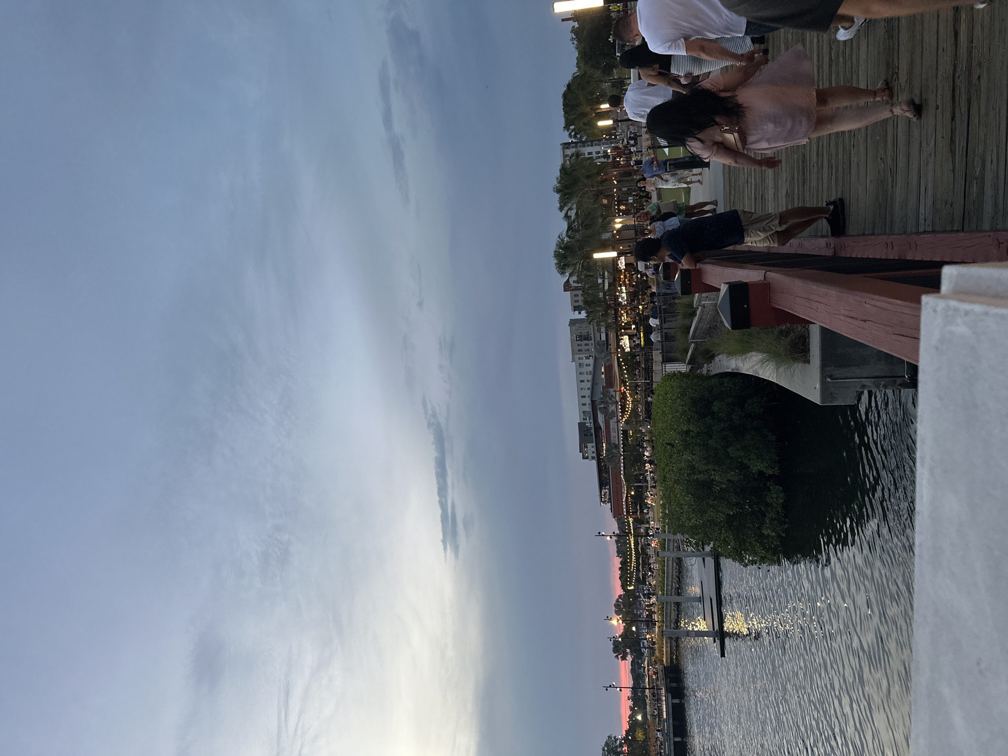 The image shows a waterfront scene in Tampa, Florida, at dusk. A group of people is gathered on a wooden boardwalk overlooking the water. Some people are standing near a red railing, looking out at the view, while others are walking along the boardwalk. Lights illuminate the buildings and the waterfront area. The sky is a mix of blue and gray with hints of pink from the setting sun. The water reflects the lights, creating shimmering patterns. A small green area with trees is visible adjacent to the water.