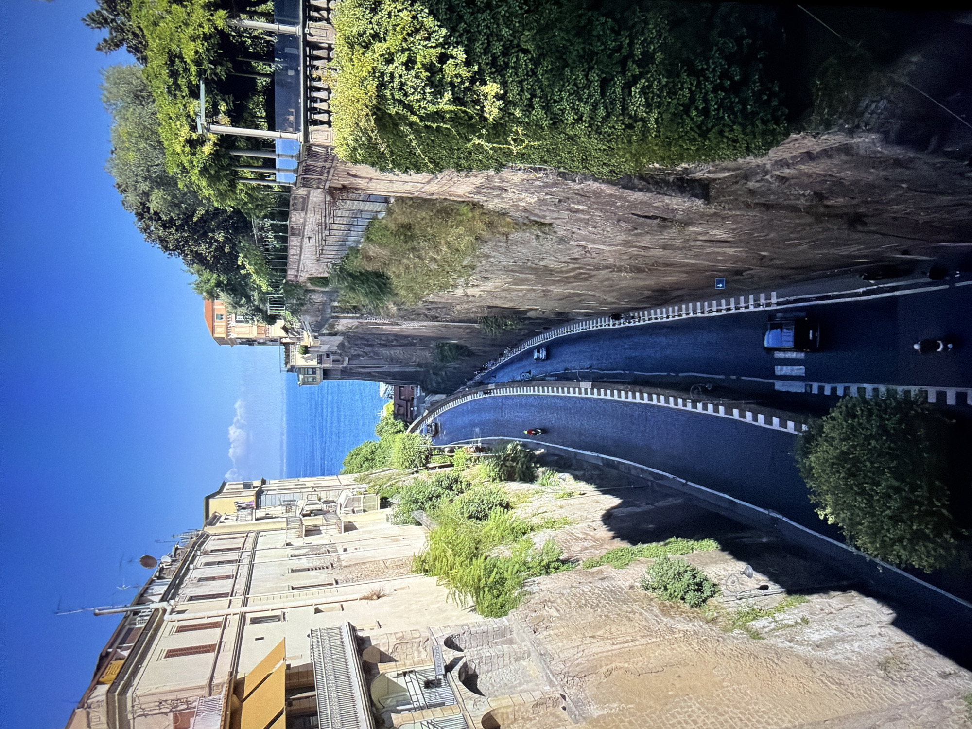 The image, correctly oriented, displays a high-angle view of a coastal town or city nestled into steep cliffs under a clear, bright blue sky. A winding, asphalt road descends from the foreground towards the background, eventually meeting the deep blue sea.
On the left side of the road, multi-story buildings with light-colored facades, balconies, and windows are visible, appearing to be integrated into the cliff face. Lush green vegetation, including trees and dense ivy, covers parts of the cliffs and complements the architecture.
The right side of the image features a dramatic, vegetated cliff, heavily covered in green foliage and trees at its summit. A promenade or walkway with decorative pillars and railings is discernible along the top edge of this cliff.
The road itself has clear white markings, including lane lines and roadside barriers. Vehicles are present, notably a black car and a person on a motorcycle wearing a red jacket, indicating ongoing traffic. Other faint vehicle shapes can be seen further along the road.
The time of day is sunny daylight, as evidenced by the clear sky and distinct shadows. A small white cloud floats above the horizon. A small blue arrow sign, pointing to the right, is visible on a road barrier. The scene suggests a warm, Mediterranean or Southern European location.