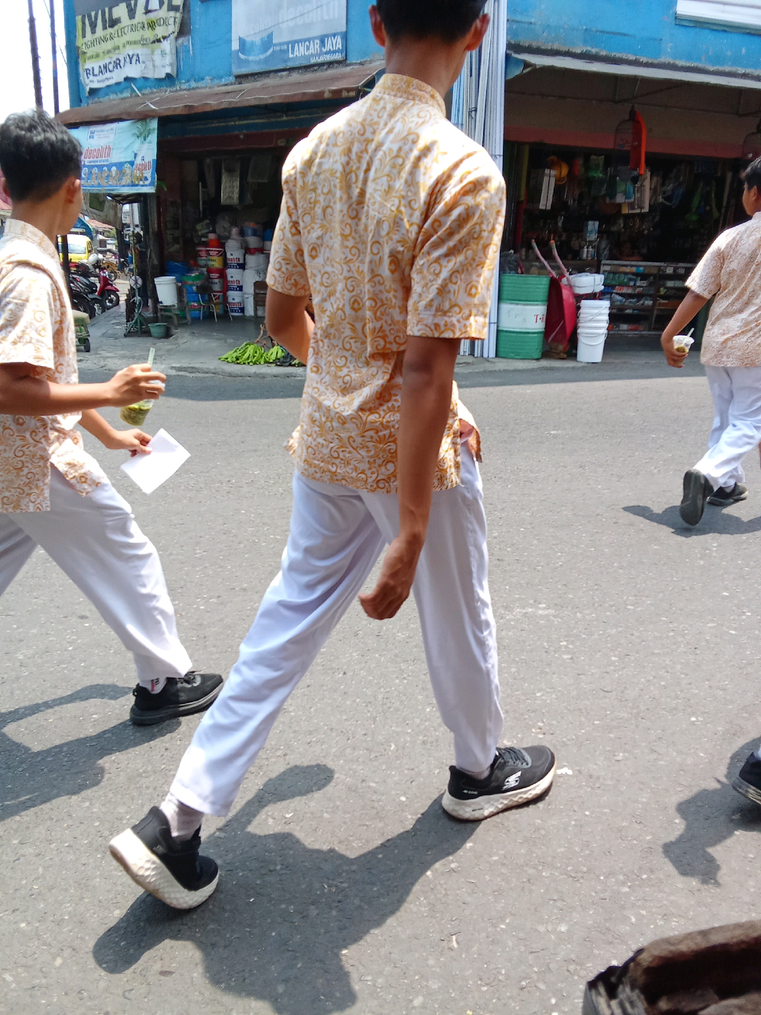 Two young males, one in the foreground and one slightly behind and to the right, are walking on a paved road in front of a storefront in Banjarnegara, Indonesia. Both are wearing light-colored, short-sleeved shirts with a repeating patterned design and white pants. The male in the foreground is carrying a clear plastic cup with a green beverage and a straw in his right hand, and a white piece of paper in his left hand. He is wearing black sneakers with white soles. The male behind him is also wearing black sneakers. The storefront appears to be a shop selling various goods, with shelves visible inside. There are also some green bananas on the ground in front of the store. The scene suggests a casual daytime activity, possibly a break or a walk through a commercial area. The environment is urban, with a sunlit street and buildings lining the road.