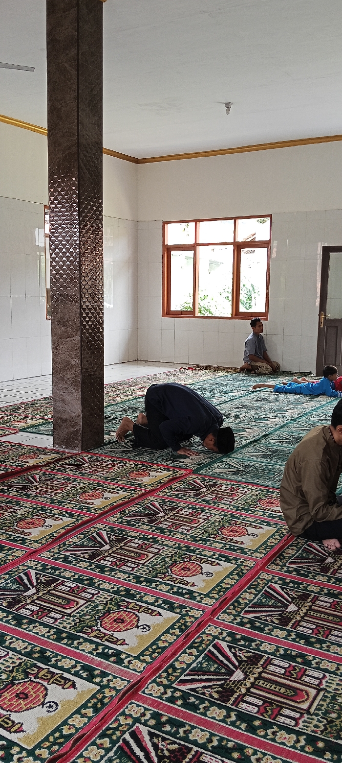 The image shows the interior of a mosque in Kecamatan Kutawaringin, Indonesia (latitude: -6.973, longitude: 107.521). The mosque is decorated with tiled walls and a patterned carpet. There are four men praying on the carpet, all facing the same direction. One of the men is standing, while the other three are kneeling and prostrating themselves in prayer. The mosque has a large window on one side of the room, and there is a door on the opposite side. The door is closed. There is a pillar near the center of the room. The men are wearing traditional Muslim clothing. There is a light fixture on the ceiling. The room has a white ceiling and white walls. The floor is covered with a patterned carpet. The carpet is green with red and gold details. The men are facing a wall in the distance. The men are all focused on their prayers.