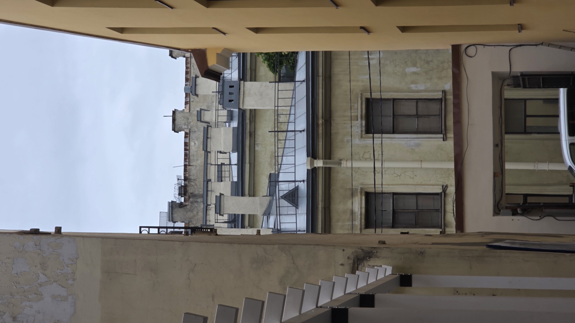 The image shows a view of an inner courtyard between buildings in Saint Petersburg, Russia. The angle is looking upwards, capturing the facades of adjacent buildings. The sky is visible at the top, indicating it's daytime. The buildings are painted in various shades of yellow and beige, with architectural details like windows, pipes, and ventilation systems. A fire escape ladder is visible on one of the buildings. The foreground includes parts of a structure, possibly a staircase or a platform, made of white and gray materials. The image conveys a sense of the enclosed space and the unique architectural features typical of Saint Petersburg's courtyard environments.