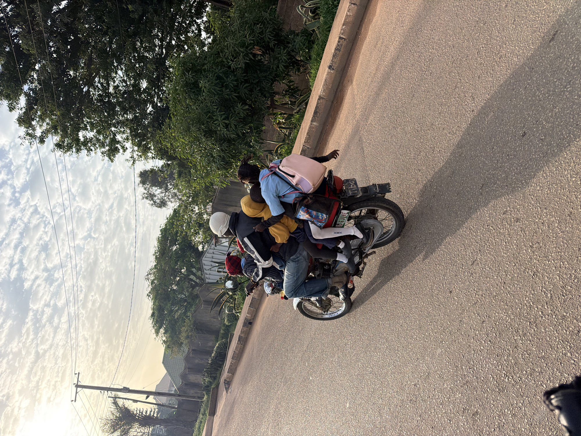 A group of five people, including at least three children, are riding on a single motorcycle on a paved road. The motorcycle is red with a license plate that reads "GUR/630YE". The driver is wearing a black jacket and a white helmet, and the other passengers are wearing various colored clothing. The motorcycle is moving forward, and the passengers appear to be seated one behind another. The road is lined with a concrete barrier and vegetation. In the background, there are trees, houses, and utility poles. The sky is cloudy with the sun shining through. The scene is set in Gura Top, Nigeria.