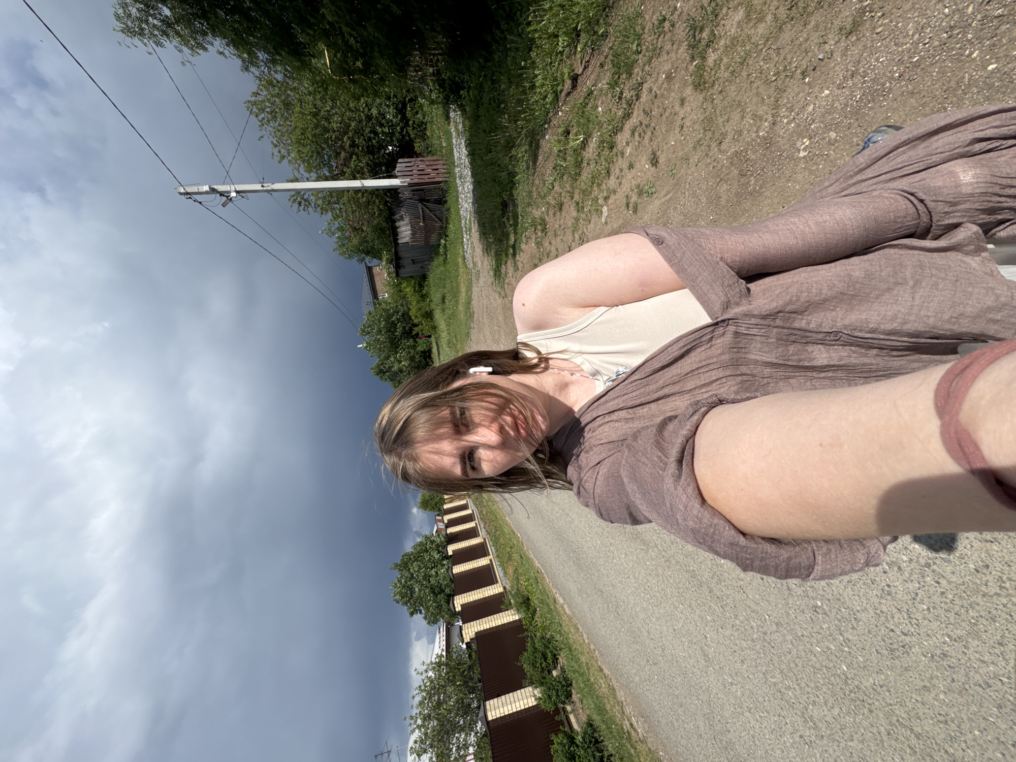 A young woman is outdoors in Горный, Russia, on a paved road between residential areas. She is wearing a brown, button-down shirt over a beige tank top, and earbuds. A fence runs along the road behind her. The sky is overcast with dark clouds. Power lines are visible in the distance.