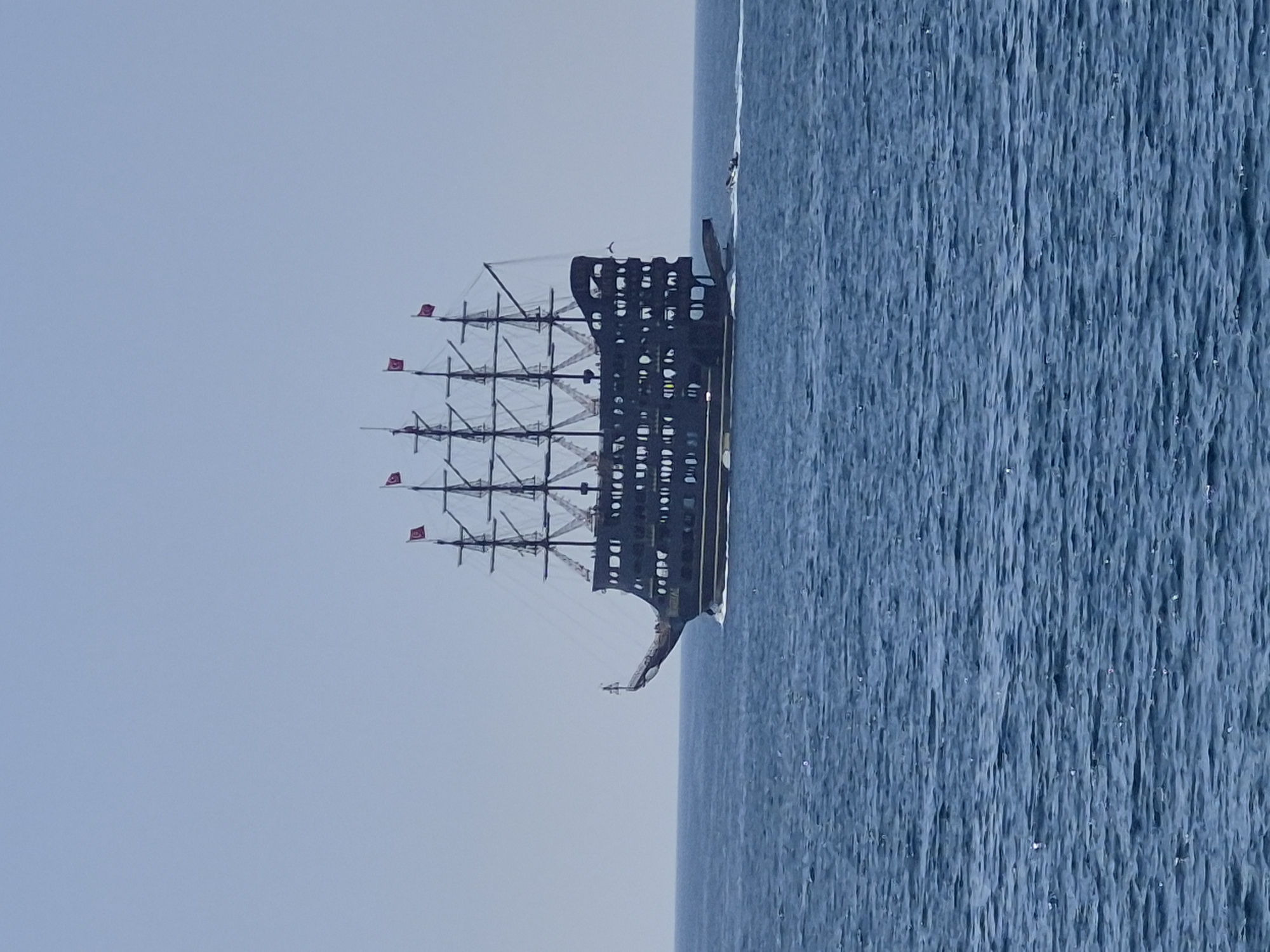 A black pirate ship with red Turkish flags sails on the water near Alanya, Turkey. The sea is blue, and the sky is a light blue.